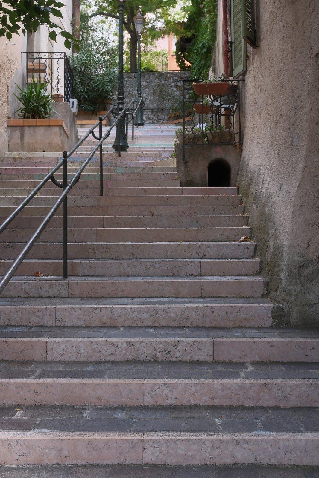 Photo verticale. Un escalier de ville avec une double rampe au centre, qui passe entre des maisons. Sur la droite, sous un palier de porte d’une des maisons, une ouverture comme une mini porte arrondie en haut.