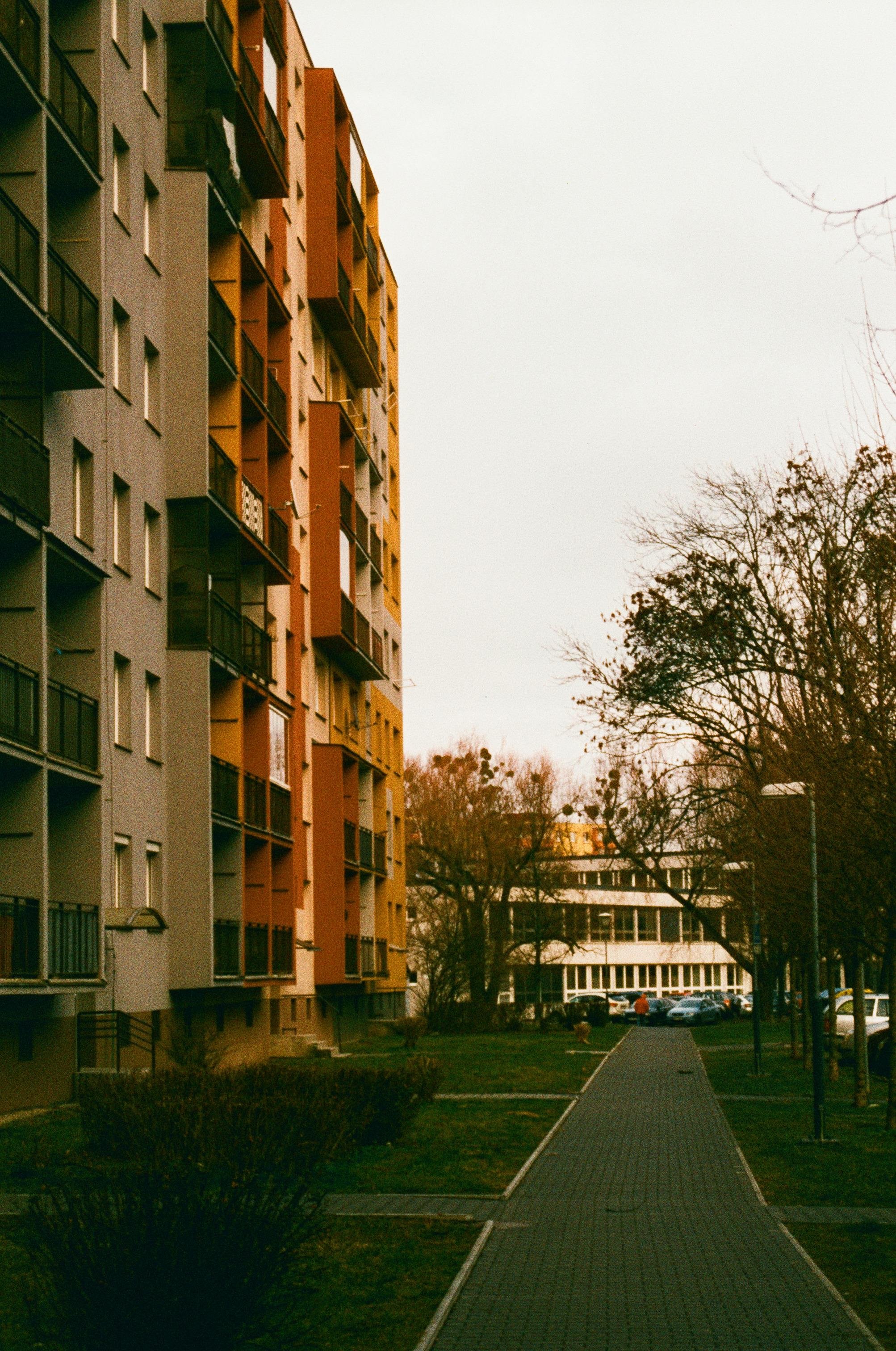 The image shows an urban scene with a focus on a multistory residential building. The building is painted in varying shades, including gray and orange. There is a pathway in the foreground leading towards other buildings in the background, and trees line the pathway with some cars and people visible in the distance. The overall atmosphere seems to be overcast.