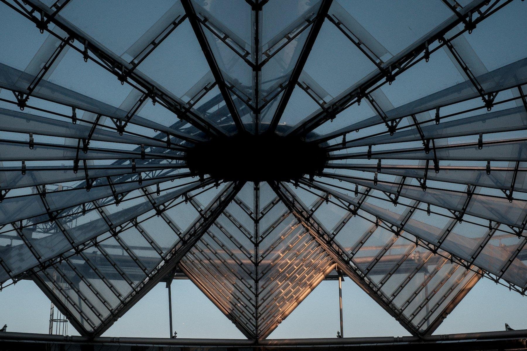 Photo horizontale. Un morceau d’une grande verrière circulaire dans une forme faisant penser à un éventail circulaire ouvert, sous un ciel de soleil couchant, une touche orangé au centre-bas de l’image
