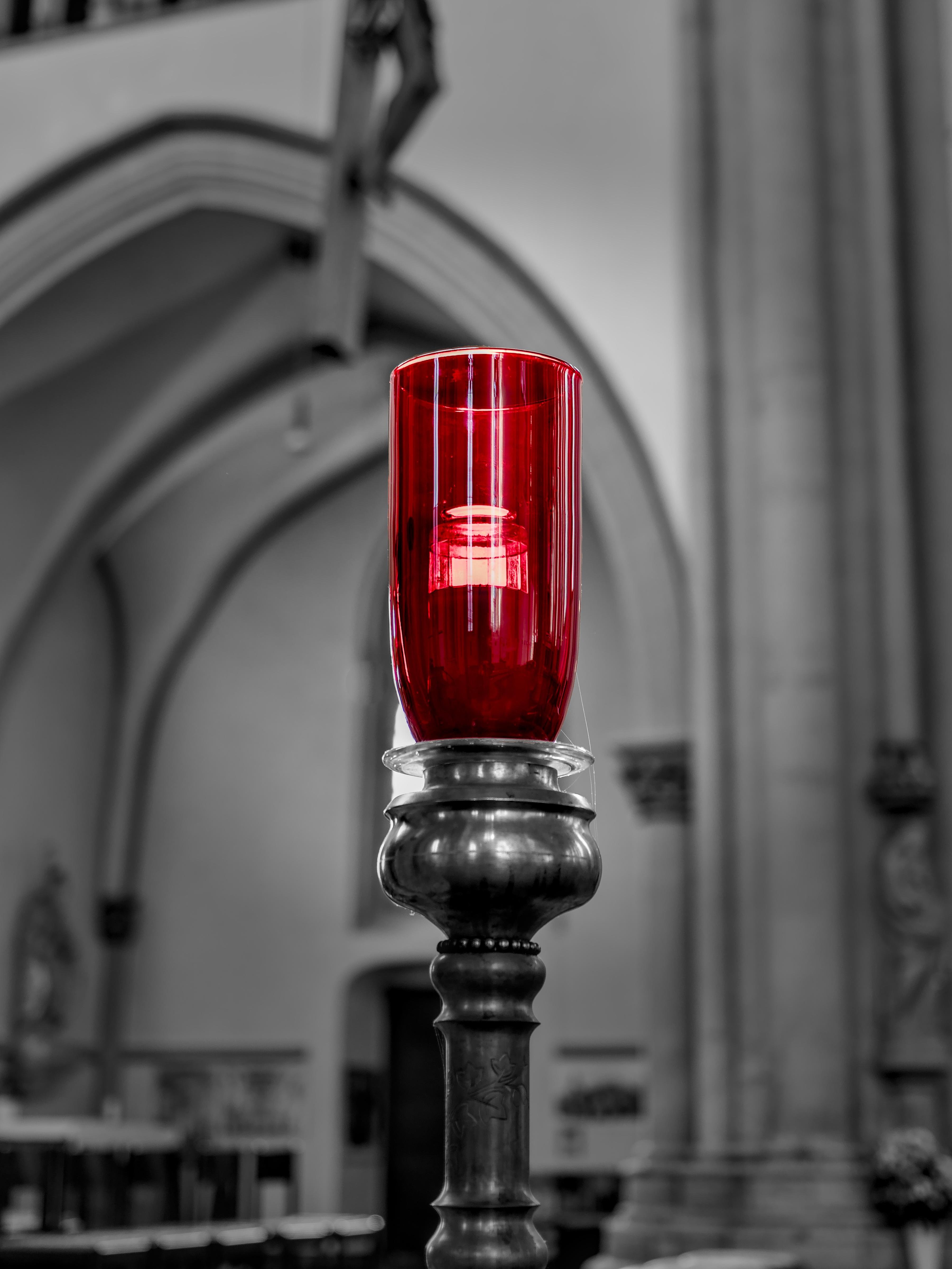 A red glass votive candle holder, illuminated by the light coming through a church window in the background, is placed on a dark metal stand and centered in the foreground against a blurred black and white church interior with arches and columns. 
### 
Roter Glas-Votivkerzenhalter, der vom einfallenden Licht durch ein Kirchenfenster im Hintergrund erleuchtet wird, auf einem dunklen Metallständer, im Vordergrund gegen ein unscharfes schwarz-weißes Kircheninneres mit Bögen und Säulen zentriert.