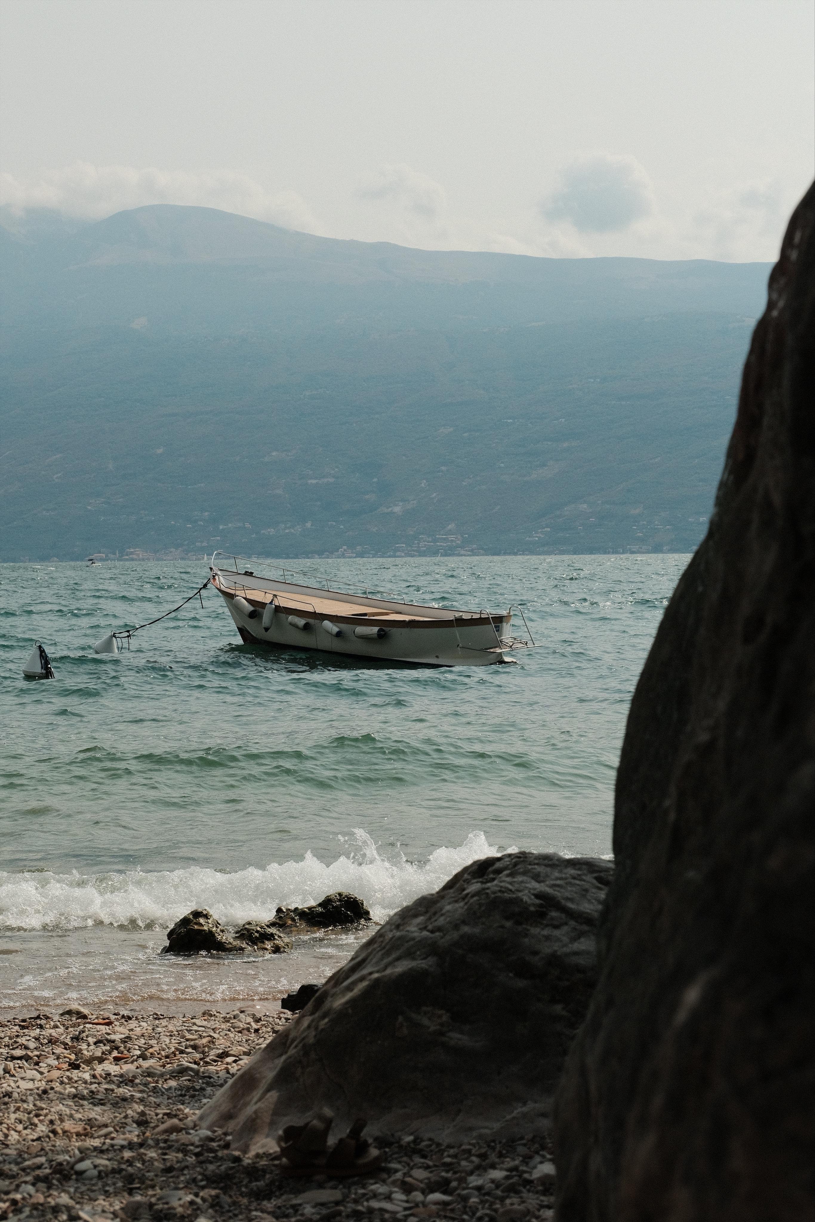 Small boat anchored on choppy water near a pebbled shoreline, with waves breaking on rocks in the foreground and hazy mountains under a cloudy sky in the background.