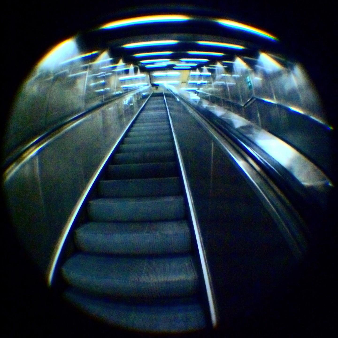 This image shows an escalator in motion, captured with a fisheye lens effect. The escalator appears to be in an indoor a subway station, with artificial lighting above.