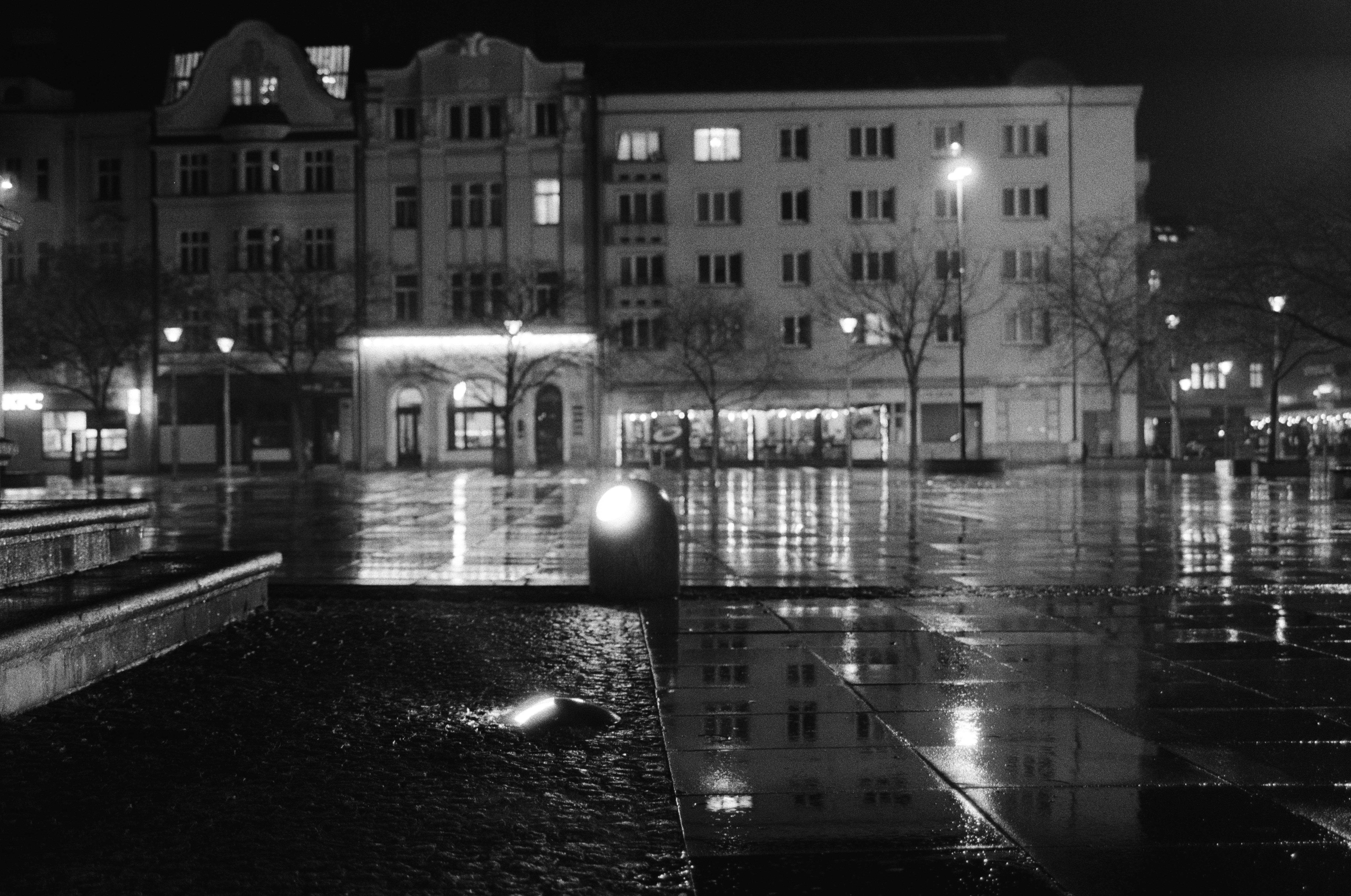The image depicts a nighttime urban scene in black and white. It shows a wet, reflective plaza or public square in front of several buildings. The area is illuminated by streetlights, and the wet ground reflects the lights prominently. The buildings have multiple stories, and there are some bare trees lining the square. The overall atmosphere is calm and quiet.