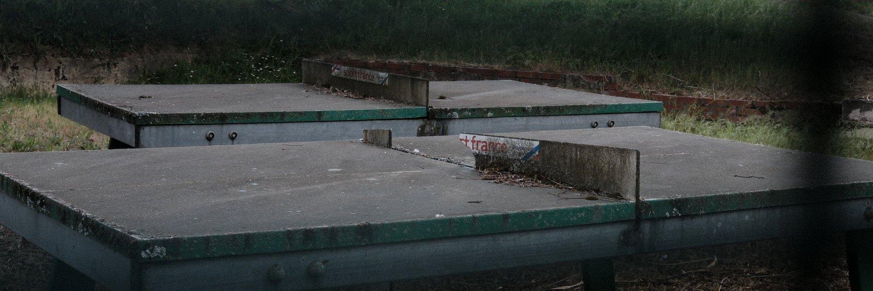 Photo horizontale au ratio très panoramique. À travers un grillage, on voit 2 vieilles tables de ping pong d’extérieur, dont le filet est en béton. Celui de la table la plus proche est cassé. Les 2 sont couvertes de crasse et de débris végétaux