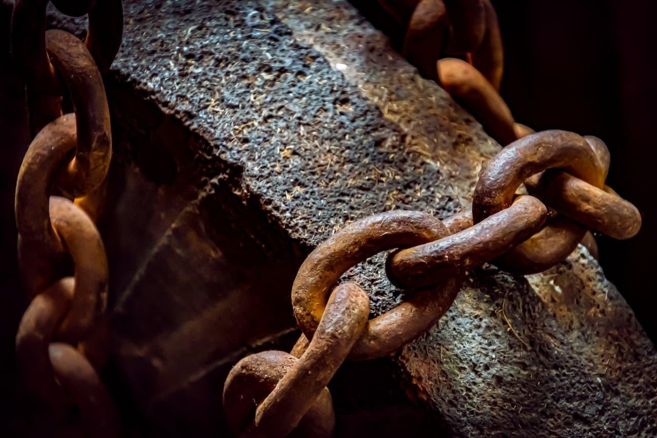 The image shows a close-up of a rusty metal chain wrapped around a rough, textured stone block. The scene conveys a sense of strength and age, with the weathered appearance of both the chain and the stone.