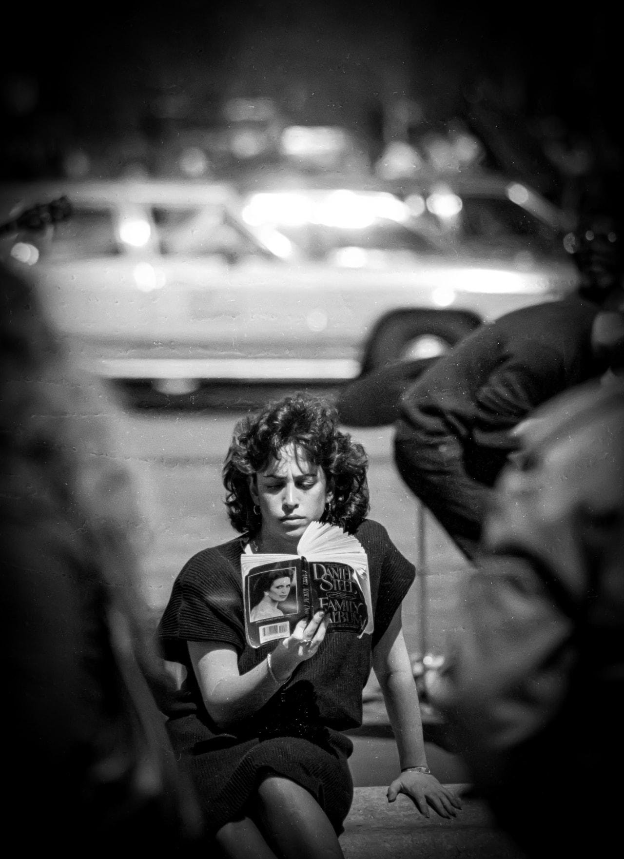 Woman sitting on a city street, focused on reading a book titled "Family Album" by Danielle Steel, with a blurred background featuring a parked vintage car. The photo is in black and white, highlighting her dark hair and the book's cover. Pedestrians are visible in the foreground and background, adding depth to the scene.
