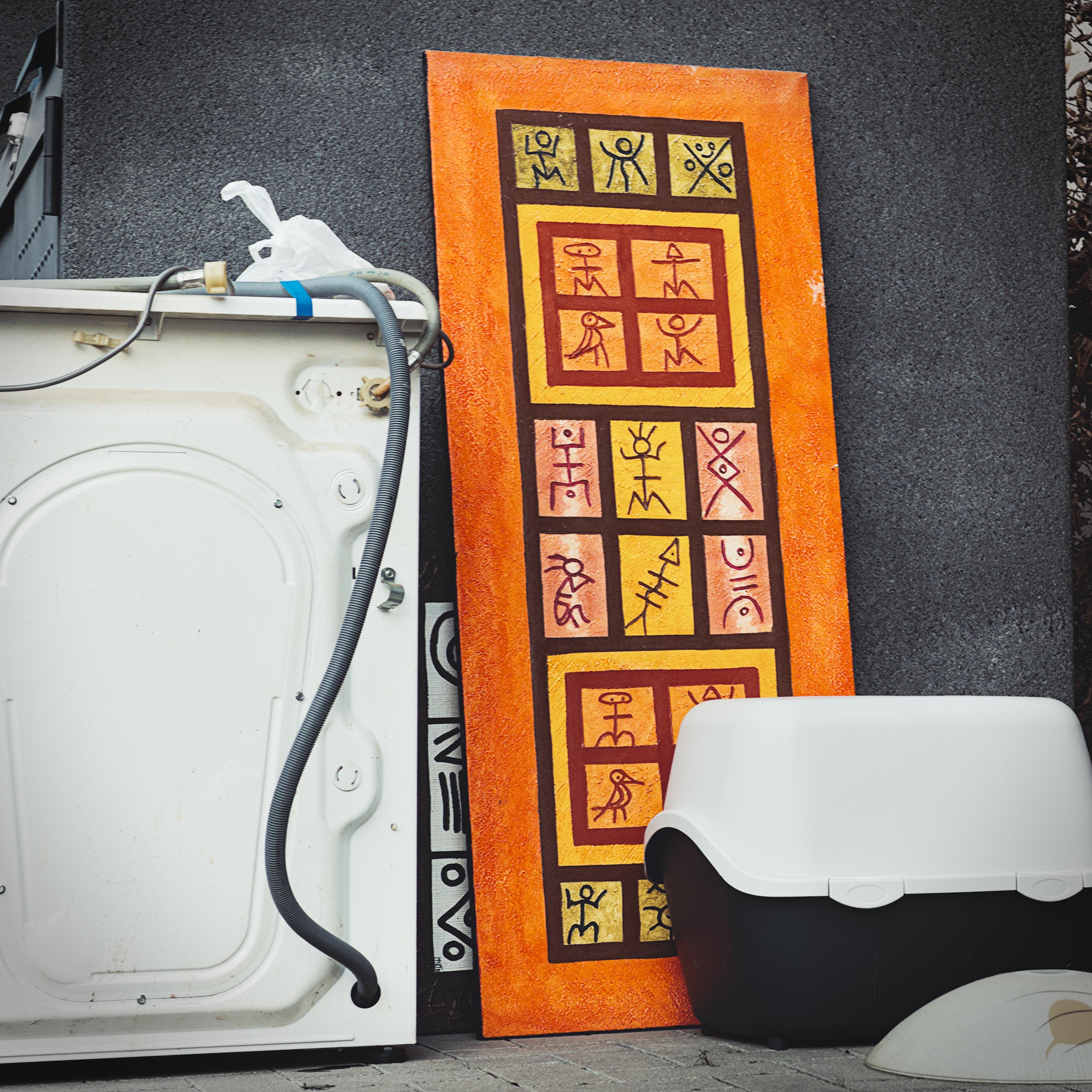 A white household appliance with exposed wires stands next to a decorative panel featuring geometric figures and symbols in red and yellow against an orange background. A black and white rectangular object, resembling a pet litter box, is in front of the panel. The scene is set against a gray wall. ### 
Ein weißes Haushaltsgerät mit freiliegenden Drähten steht neben einer dekorativen Platte mit geometrischen Figuren und Symbolen in Rot und Gelb vor orangefarbenem Hintergrund. Ein schwarz-weißes rechteckiges Objekt, das einer Katzentoilette ähnelt, befindet sich vor der Platte. Die Szene ist gegen eine graue Wand gesetzt. 