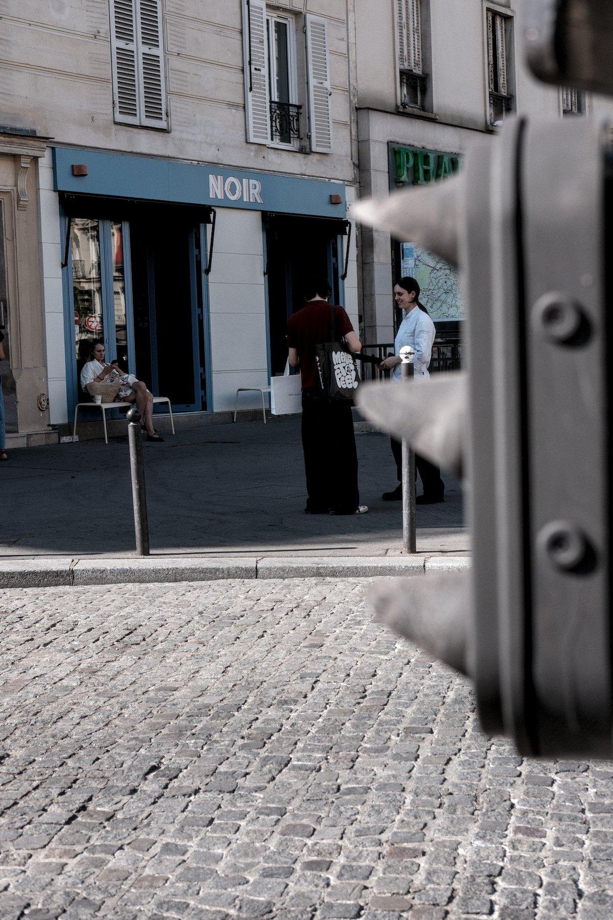 Photo verticale. Au premier plan, le petit feu tricolore du bas, flou, derrière, une route pavée au soleil, puis une place piétonne à l’ombre. Une jeune femme en t-shirt et pantalon, avec un sac de shopping blanc accroché au coude gauche et un sac tote noir aux grosse lettres blanches, rencontre une amie habillée d’une tenue blanche de travail en cuisine. Elle est cadrée de sorte à être entre 2 pare-soleil du feu tricolore. Derrière, un commerce à la devanture bleu et blanche, qui s’appelle « NOIR »