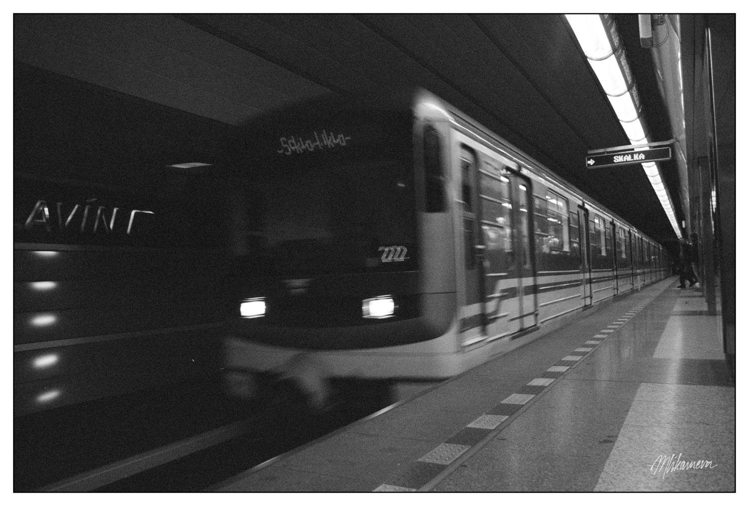 A black and white photograph of a subway train in motion at a station. The train is moving from left to right, with blurred details indicating speed. Overhead lights illuminate the platform, and a sign displaying the station name hangs above on the right. A few people are visible in the distance on the platform.