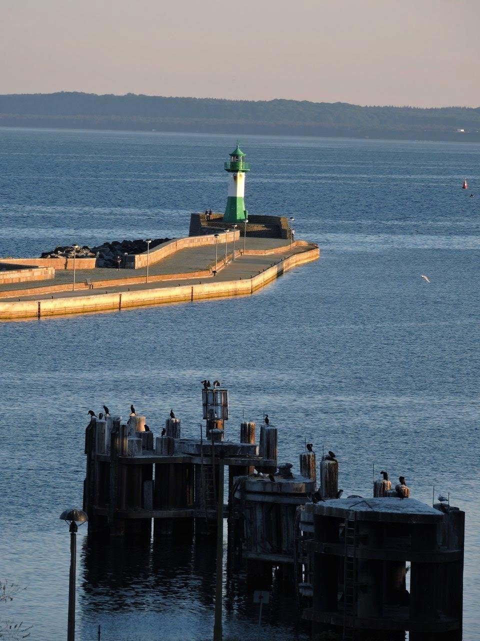 Green and white lighthouse on a pier extending into a body of water, with mountains visible on the horizon. Birds perch on wooden pilings in the foreground. The scene is illuminated by soft, late afternoon sunlight.