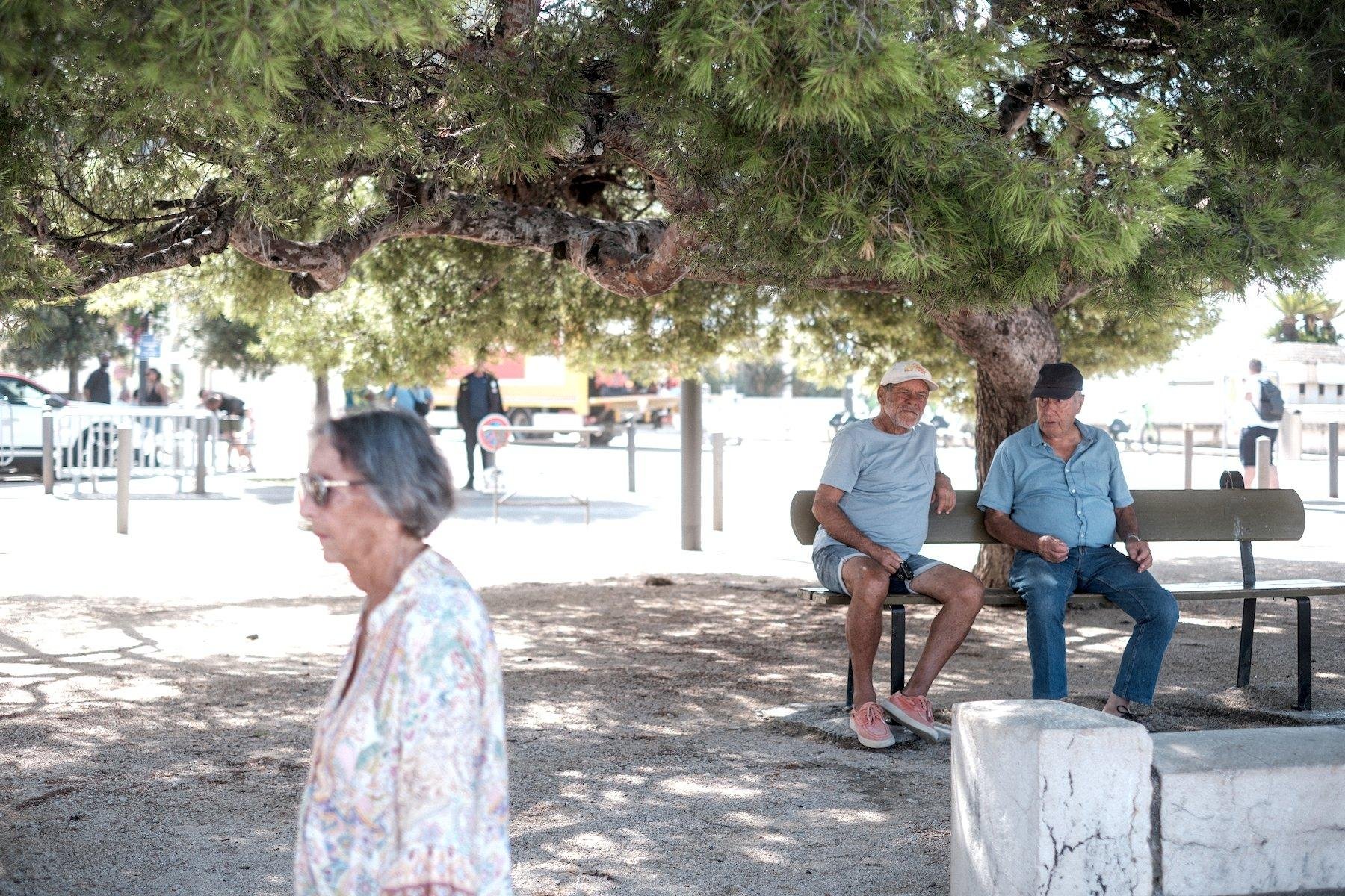 Photo horizontale. Au premier plan, à gauche, une dame aux cheveux courts et gris, lunettes de soleil et un haut blanc à motifs colorés marche vers la gauche. Au second plan, à droite, 2 hommes assis sur un banc publique, en train de discuter, à l’ombre d’un grand arbre poussant très horizontalement