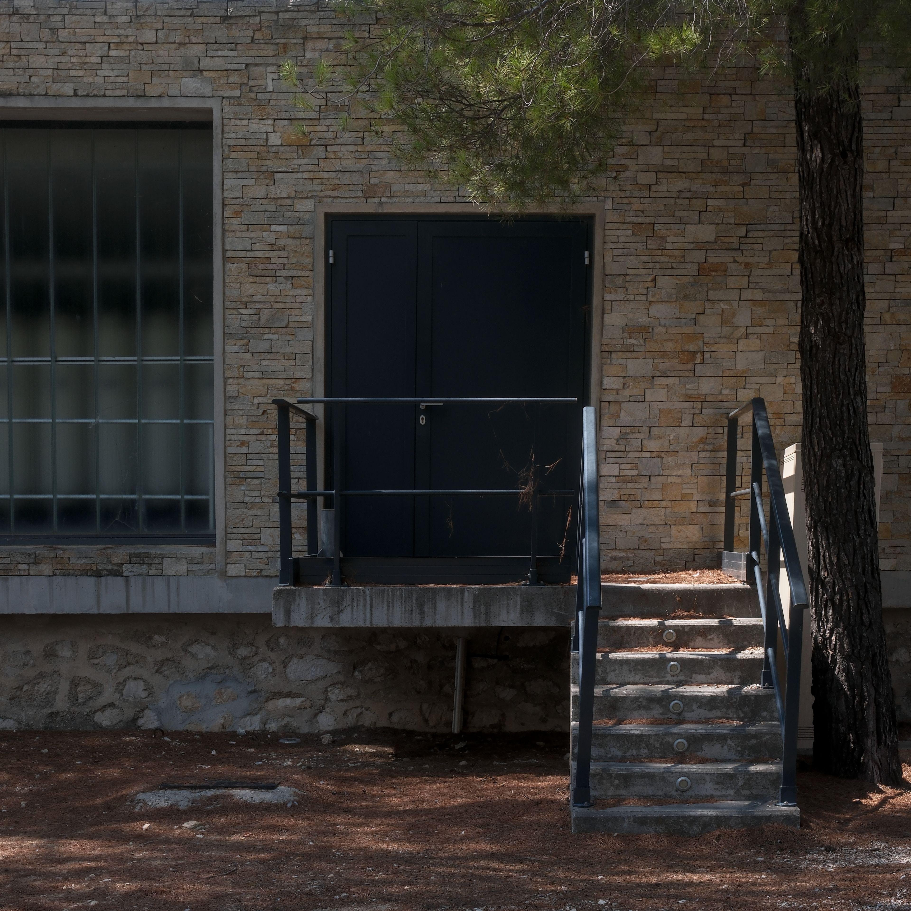 A square photo of a black door deserved by a small platform and a small 8 steps stairs. Squared fogged windows blocks can be seen on the left, starting a big window bay, and a tree close the frame at the right