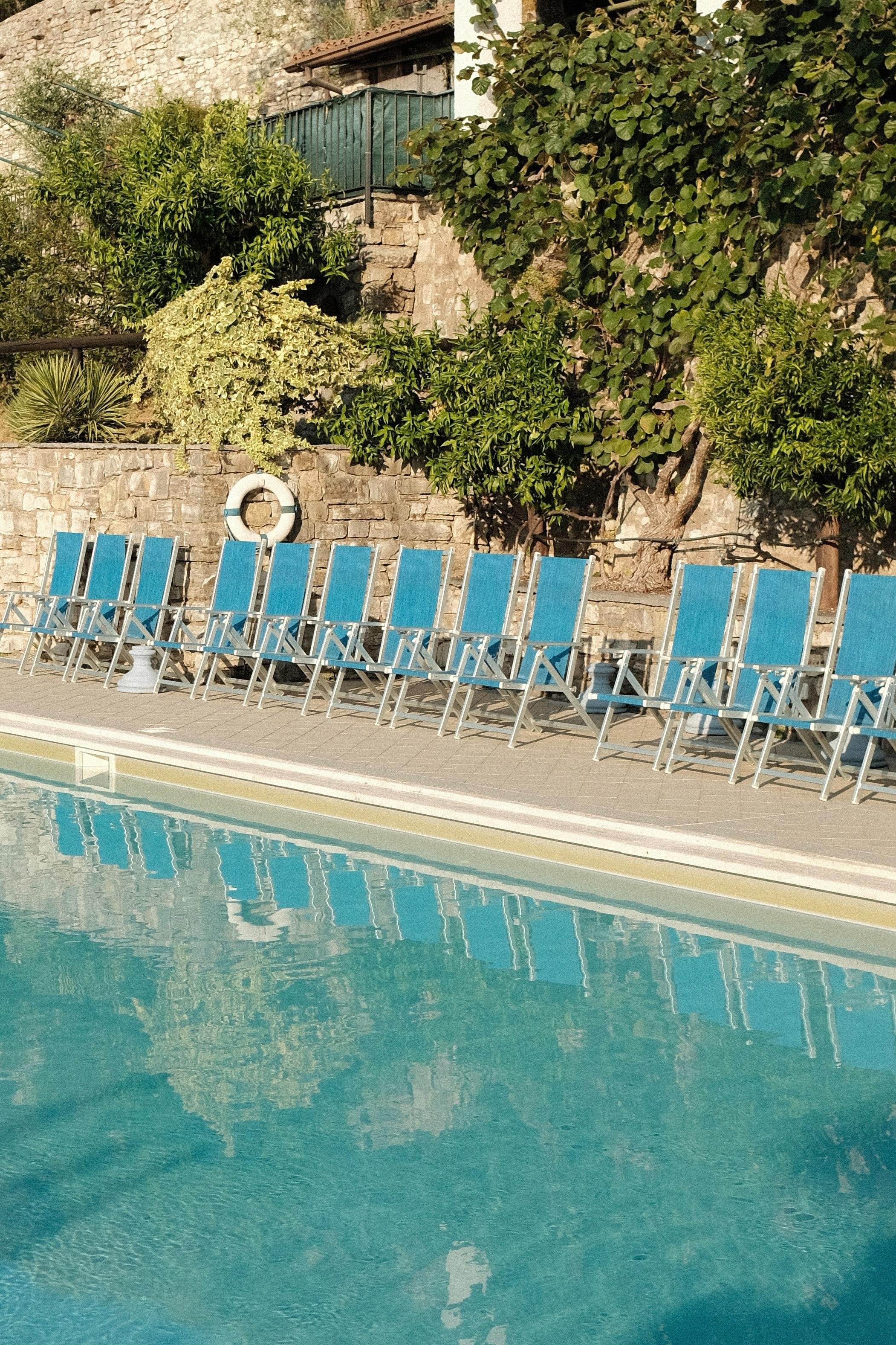 A turquoise pool with a calm, reflective surface. Several blue poolside chairs along the edge of the pool. A white life belt attached to a small stone wall behind the seats. Dense bushes on the slope over the wall.