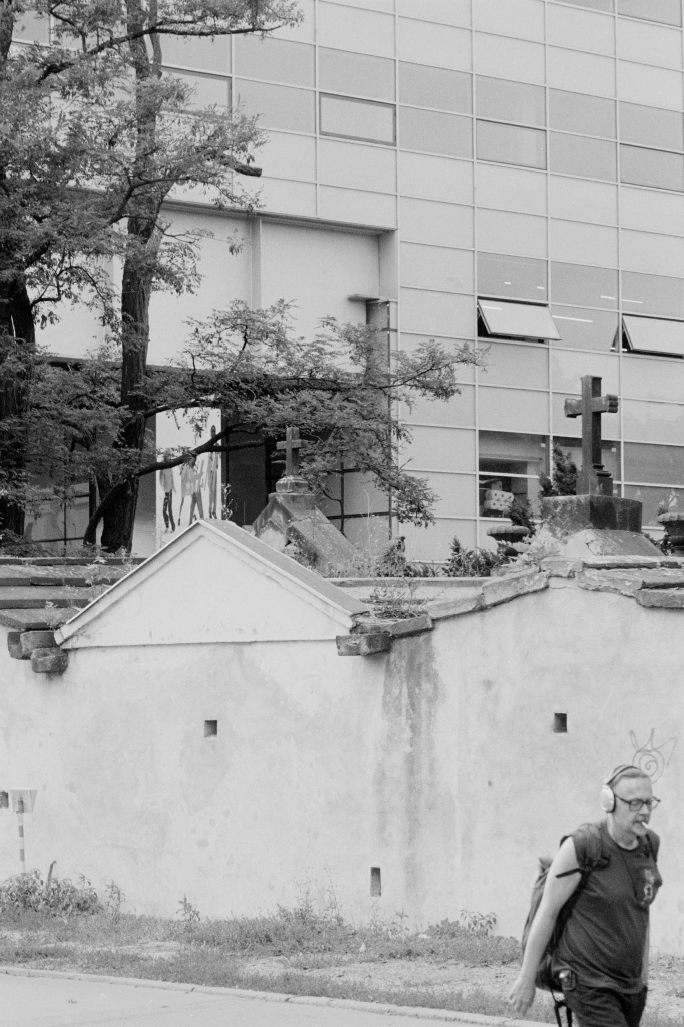 Black-and-white street scene with a man wearing headphones and a backpack walking along the bottom edge; a low plaster wall with a tiled roof and stone crosses behind him, trees rising above, and a modern glass office building with some windows propped open in the background.
