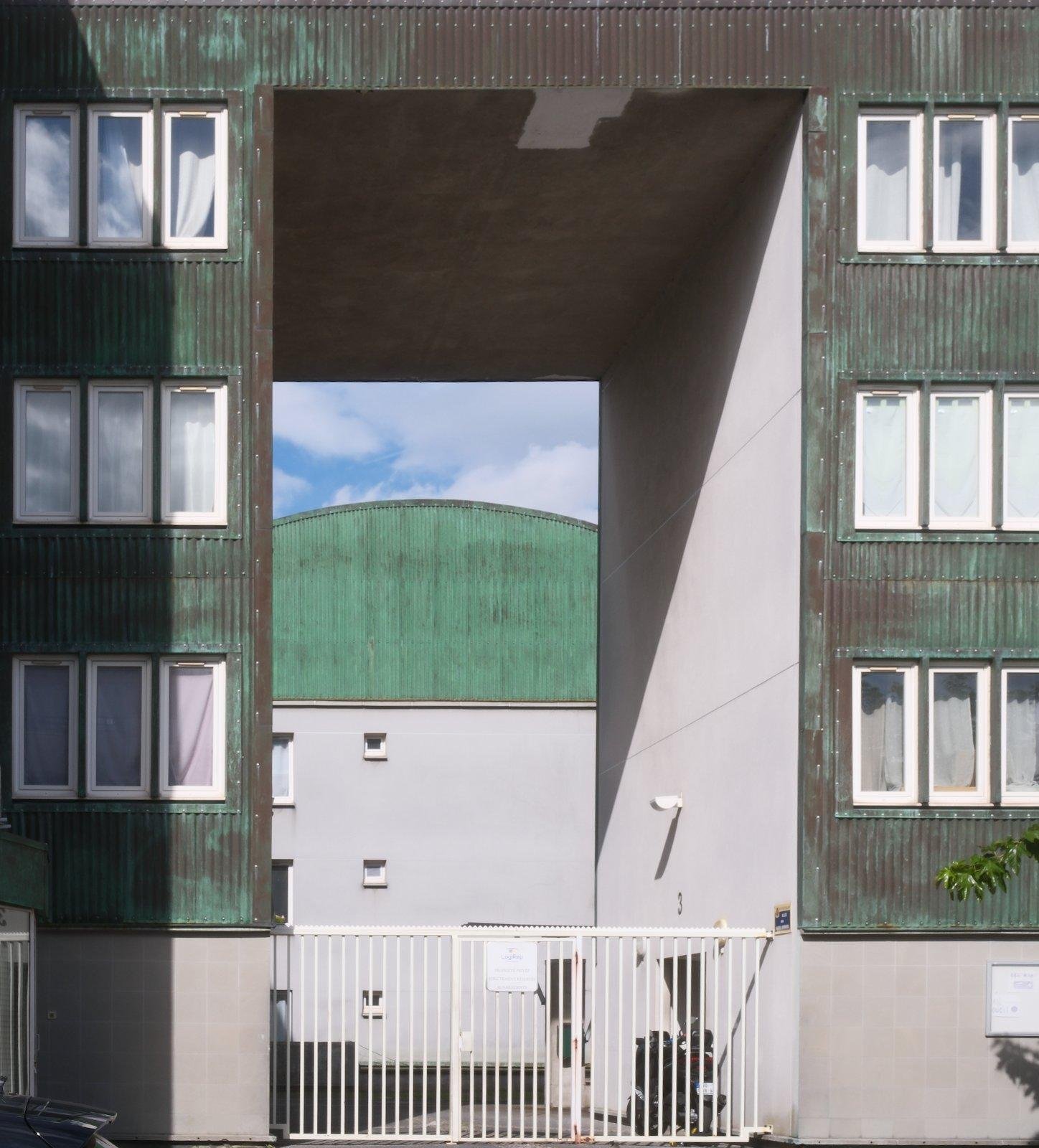Square photo. face of a building consisting of a big arche which colour is dark green with a lot of rust on it. Across the arch, we can see another warehouse, with the same green and rust on the wall.