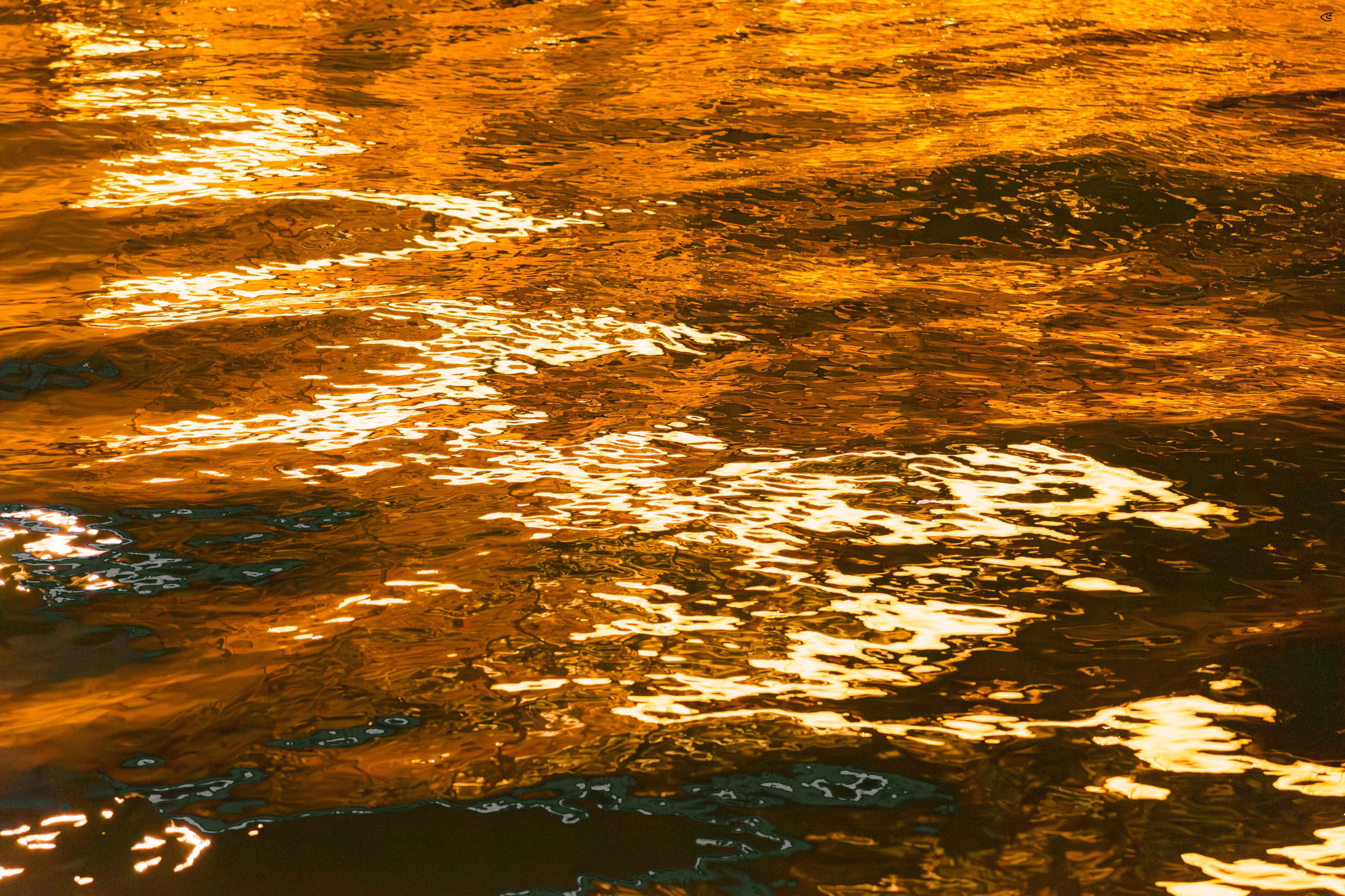 Close-up of rippling water surface with bright golden-orange reflections across most of the frame and darker shadowed areas along the bottom edge.