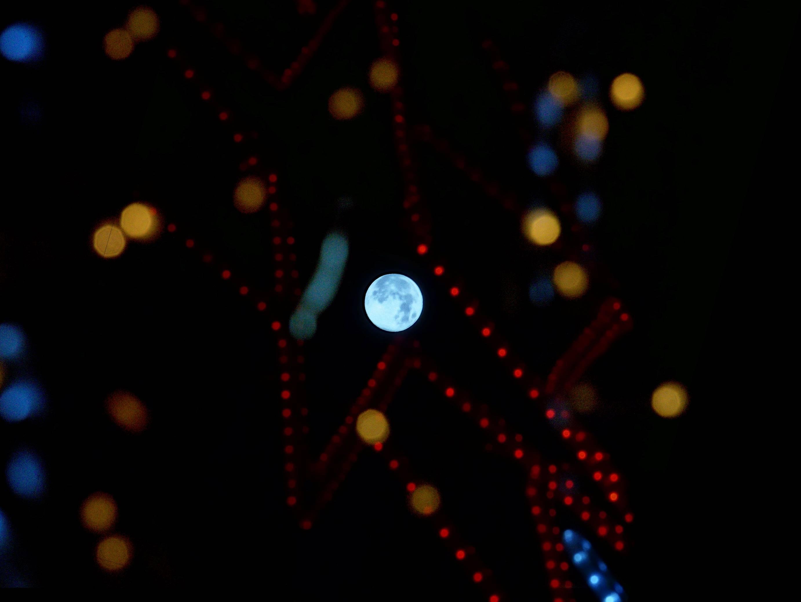 L'image montre la lune visible à travers une constellation de lumières colorées. Les lumières sont floues et apparaissent comme un bokeh rouge, jaune et bleu sur le ciel nocturne sombre. La lune est mise au point, bien en évidence au centre de l'image.
