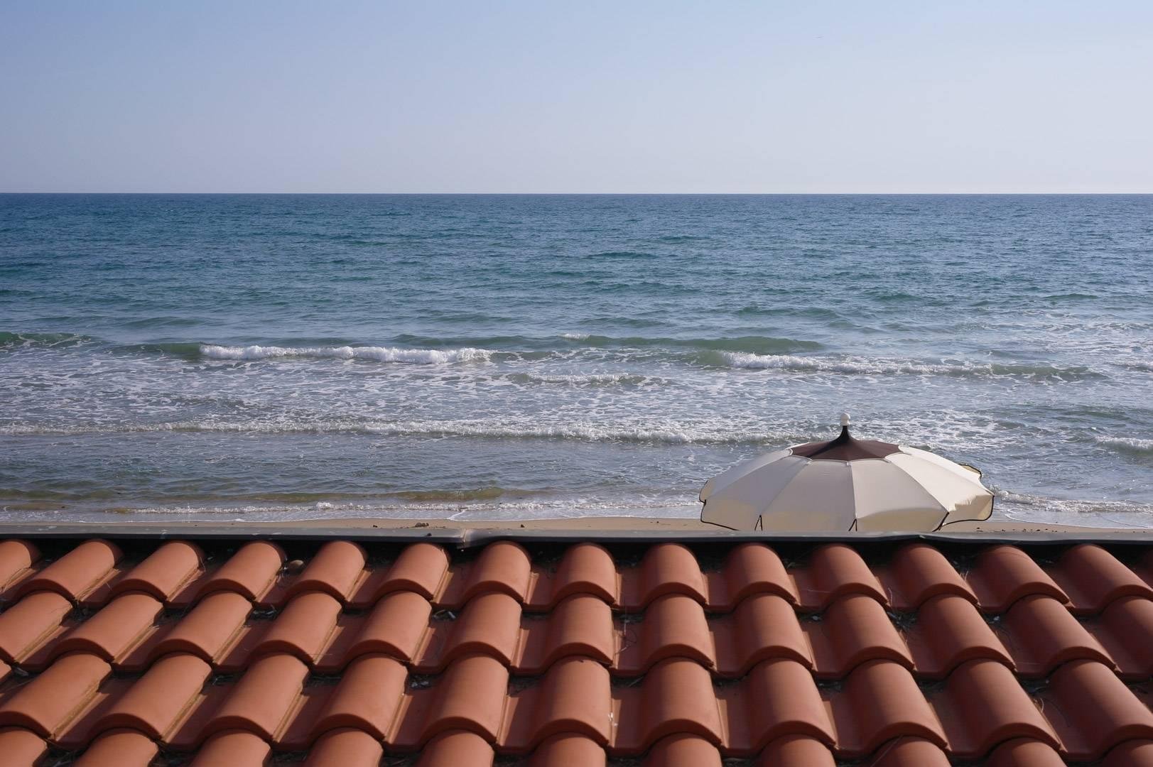 Vista del mare, col cielo limpido all'orizzonte, un ombrellone  aperto sul lato destro e un tetto di tegole nella fascia bassa della foto.