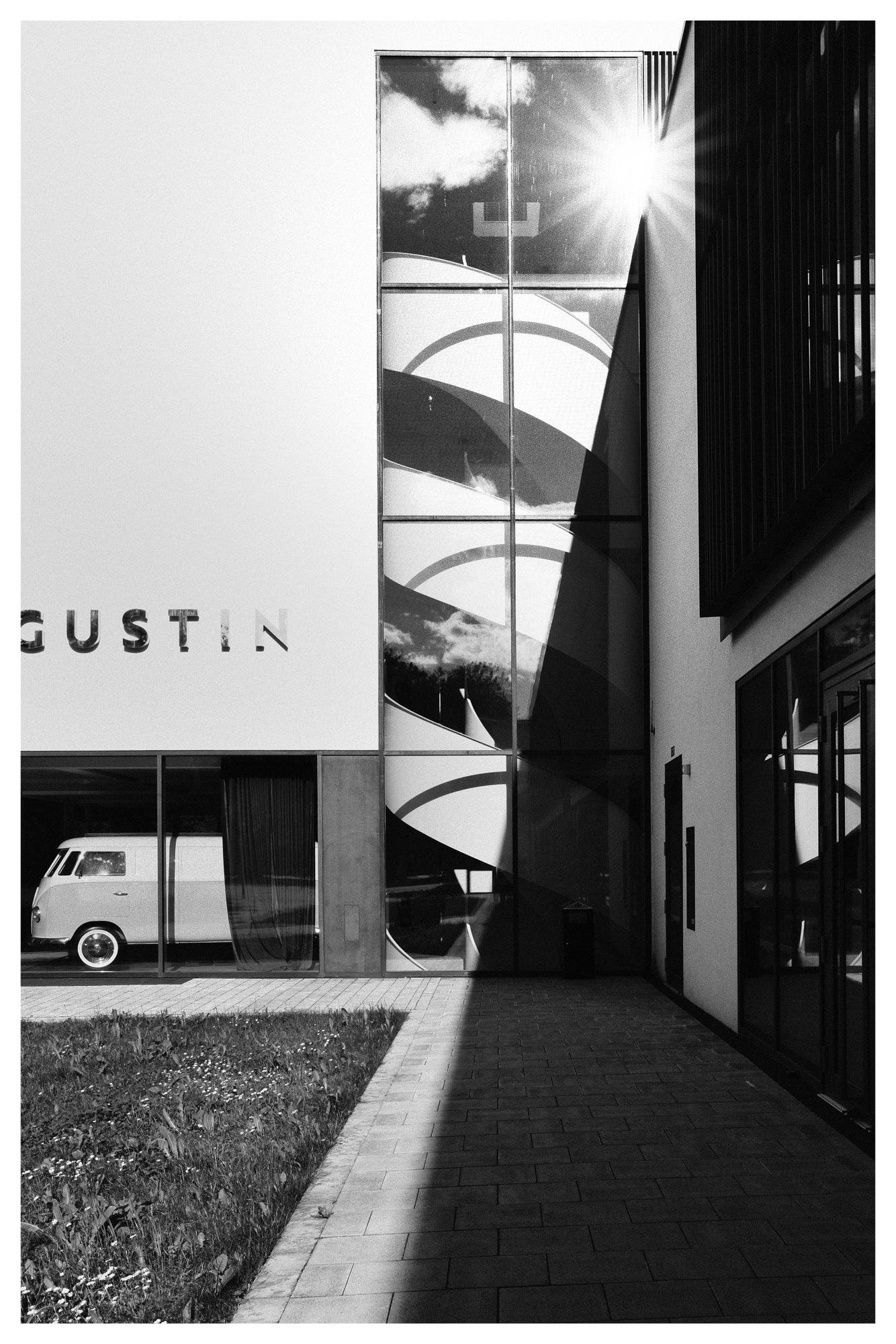 Modern building facade in black and white with a tall central glass panel reflecting clouds and showing curved graphic shapes; partial wall lettering reads “GUSTIN” on the left above a window displaying a parked van. Sun flare at the upper right, with a paved walkway and grass strip in the foreground and deep shadows along the right side near the entrance.