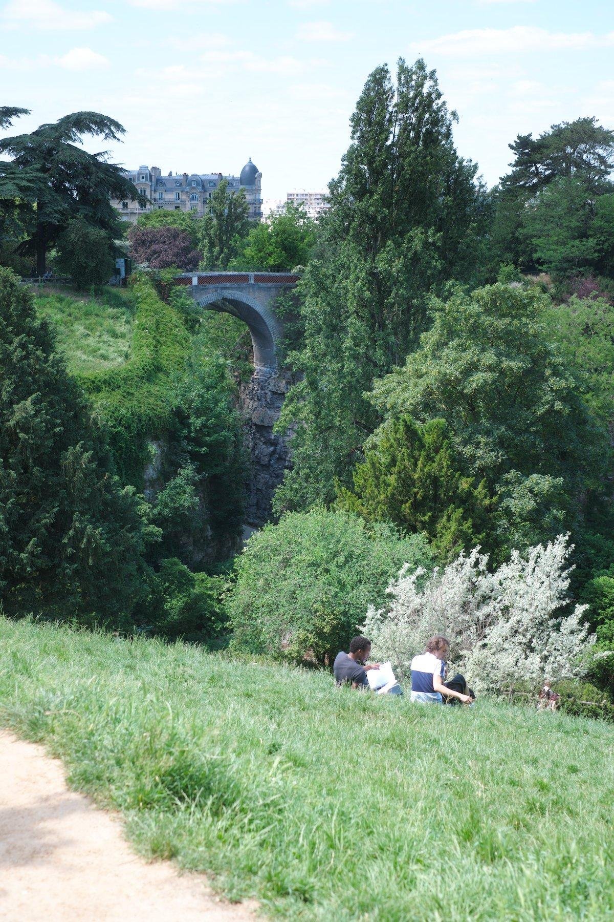 Photo verticale. Vu sur une pelouse qui est en pente descendante très raide. 2 hommes sont assis dessus. On voit un pont en pierre au fond, construit sur les bords d’une falaise. Des arbres sont présents un peu partout
