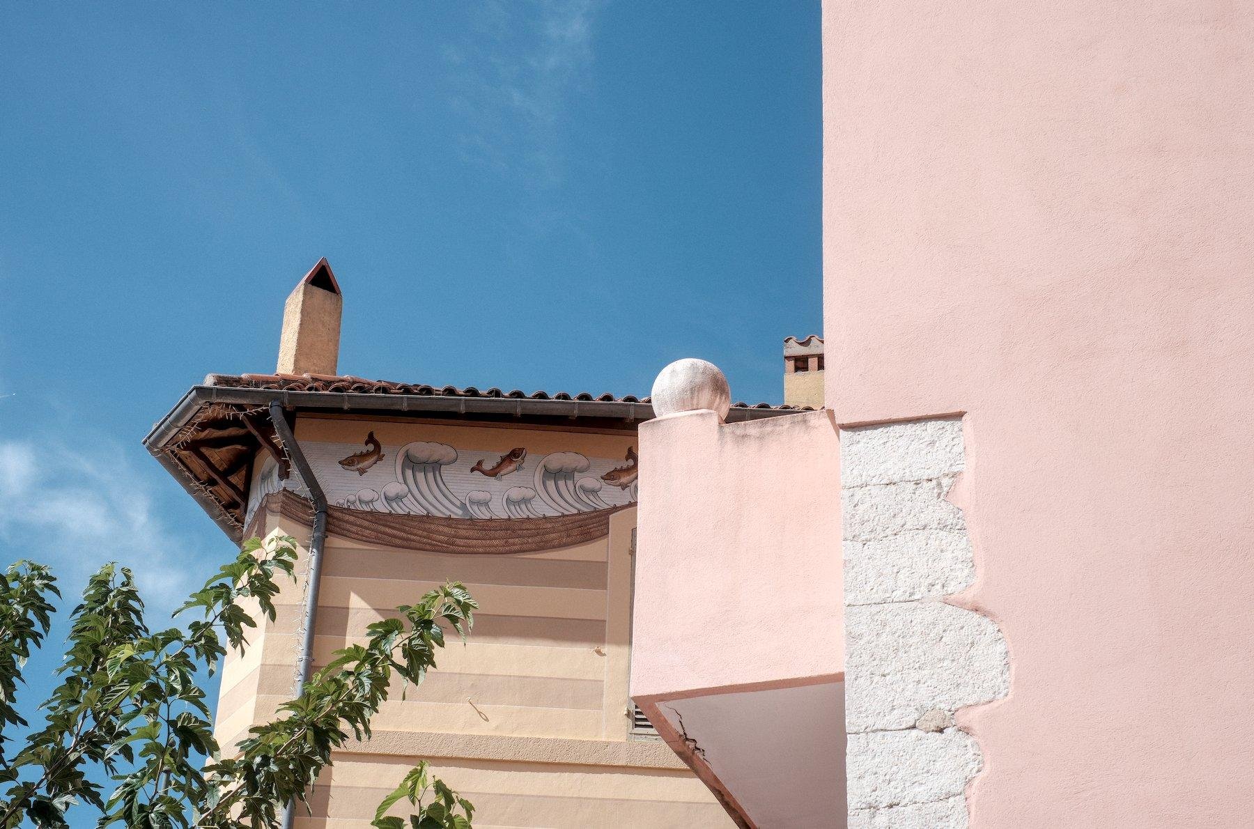 Photo horizontale. Sur la droite au premier plan, un mur rose dont une partie de la couche peinte s’est effrité dévoilant des pierres taillées blanches. Sur la gauche, le ciel bleu se dévoile avec dessous un autre bâtiment avec une fresque peinte en haut, sous le toit, représentant des poissons au dessus de vagues. En bas à gauche, des branches feuillues, vertes