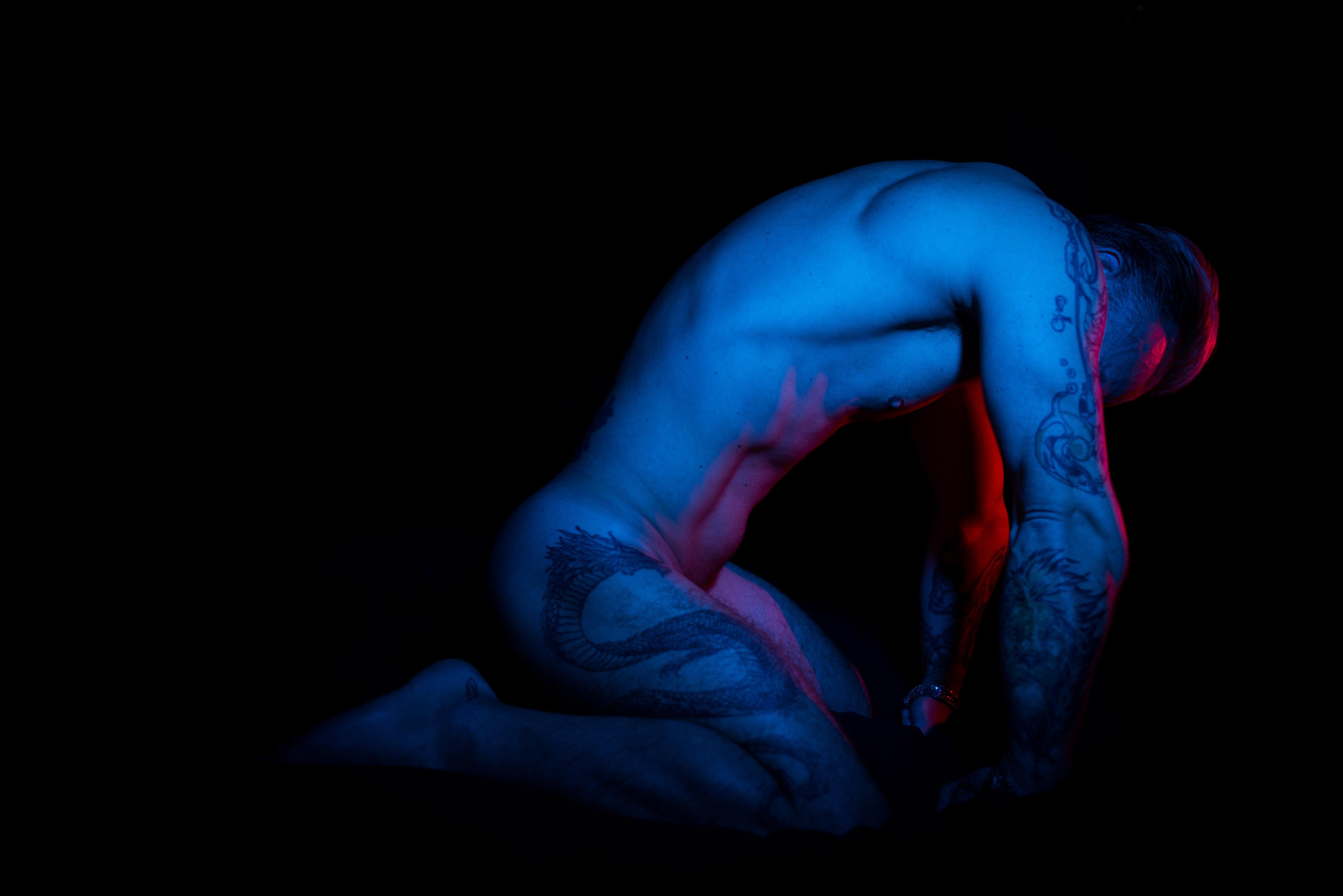 The image features a person kneeling, illuminated by colorful lighting, primarily blue and some red. The person's body is artistically lit to highlight muscular definition, and there are visible tattoos. The setting appears to be dark, enhancing the contrast created by the lighting.
