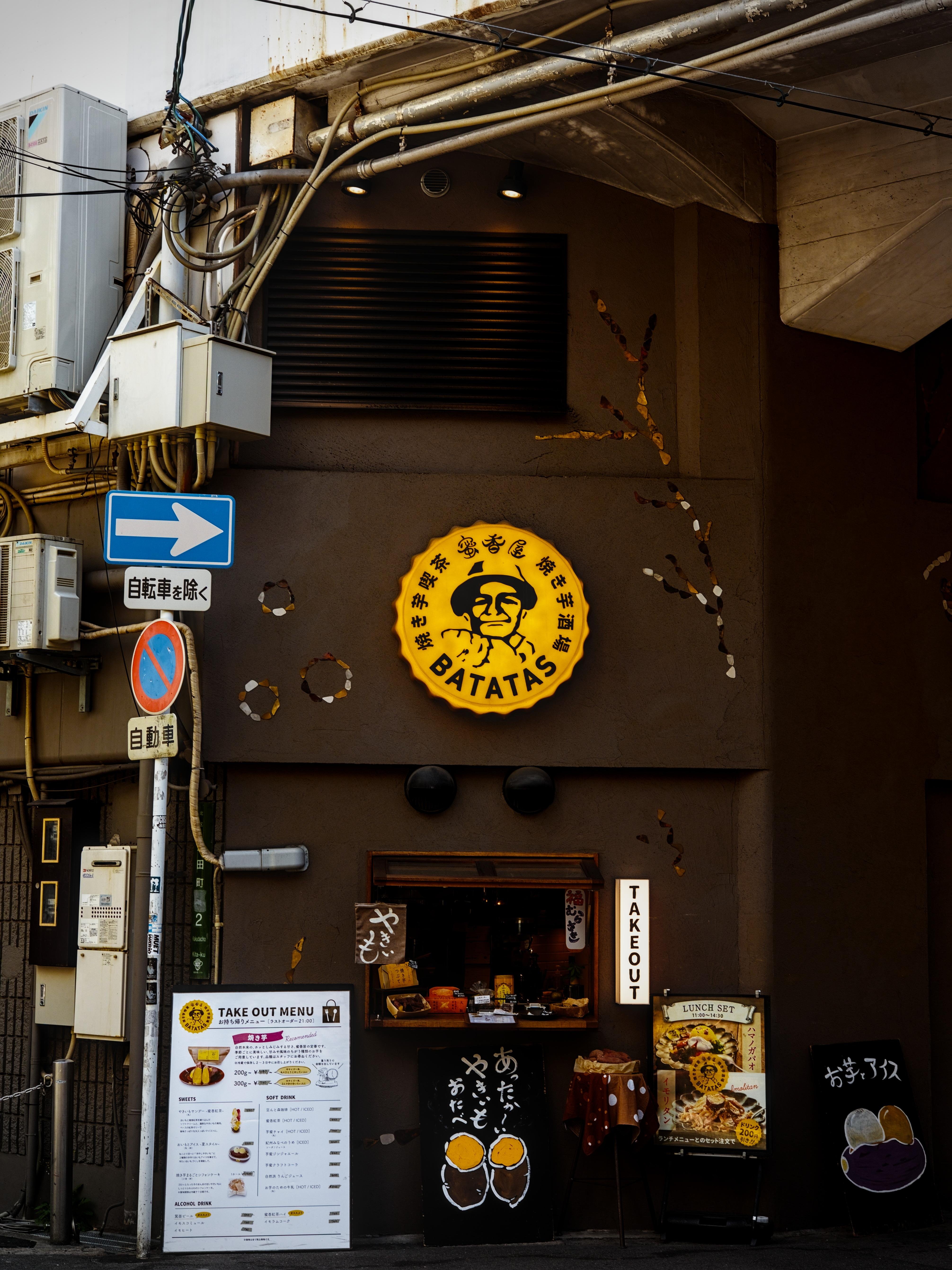 A restaurant located at the corner of two streets under a railway bridge in Umeda, Osaka. There are many signs throughout the picture: road signs, the restaurant logo, the menu, daily dish, etc.