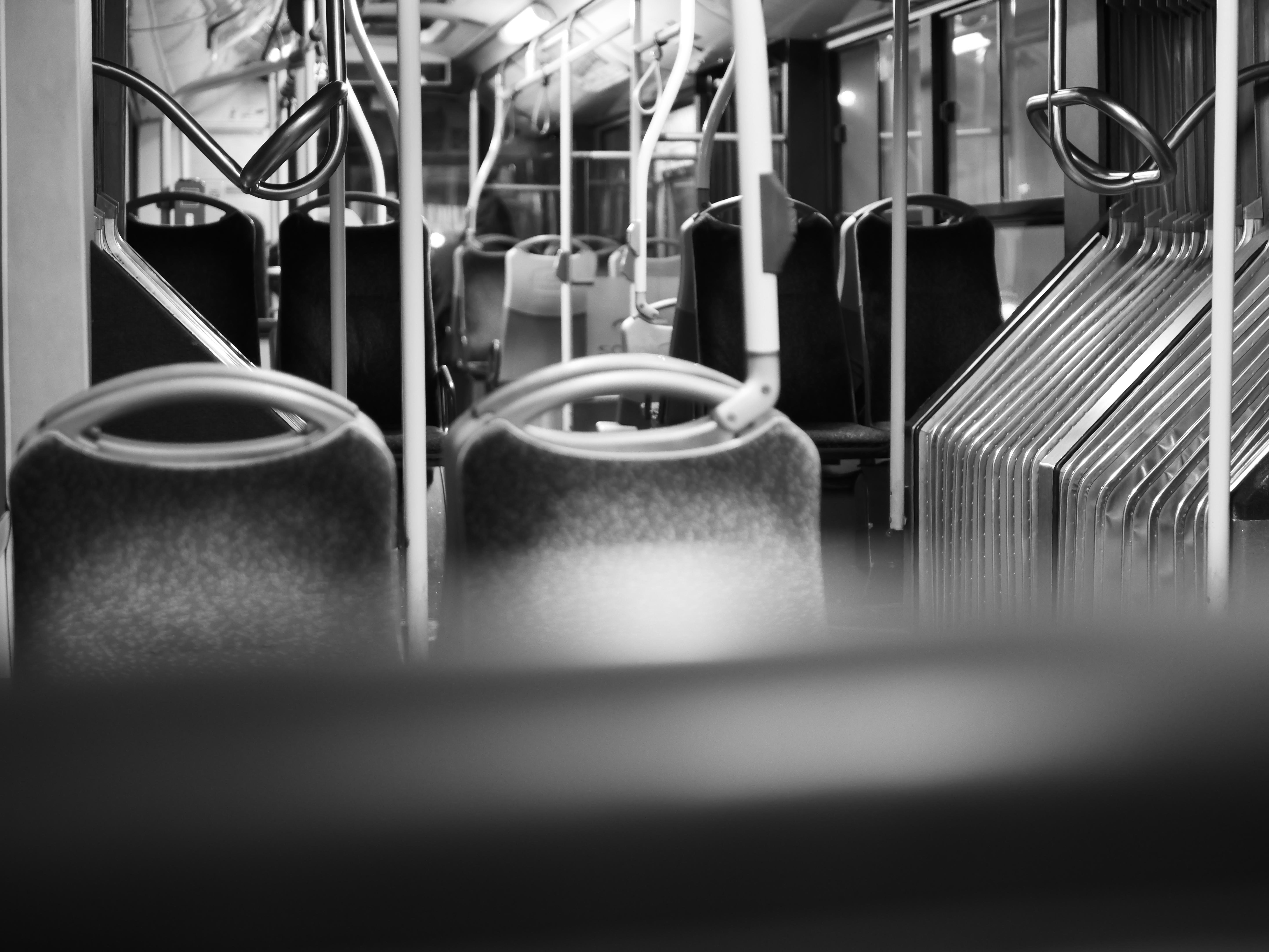 Black-and-white view inside an empty bus, looking down the aisle past fabric seat backs, with overhead hand straps and a row of stacked metal seat frames on the right.