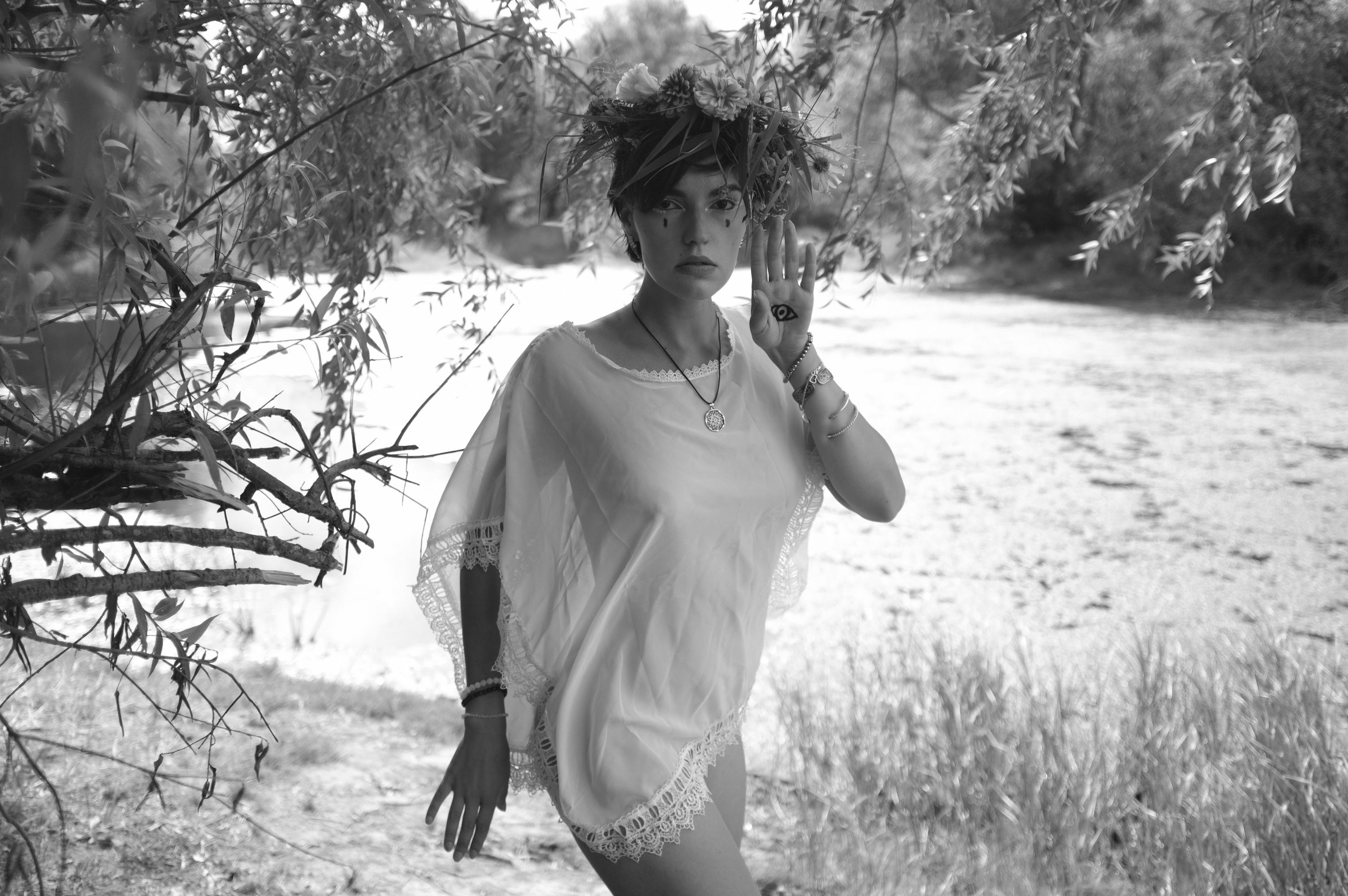Woman wearing a white lace-trimmed garment, adorned with a floral headpiece, standing near a tree with overhanging branches. She raises one hand, displaying a symbol on her palm. The background features a blurred, sunlit outdoor setting with grass and trees. Image is in black and white.
