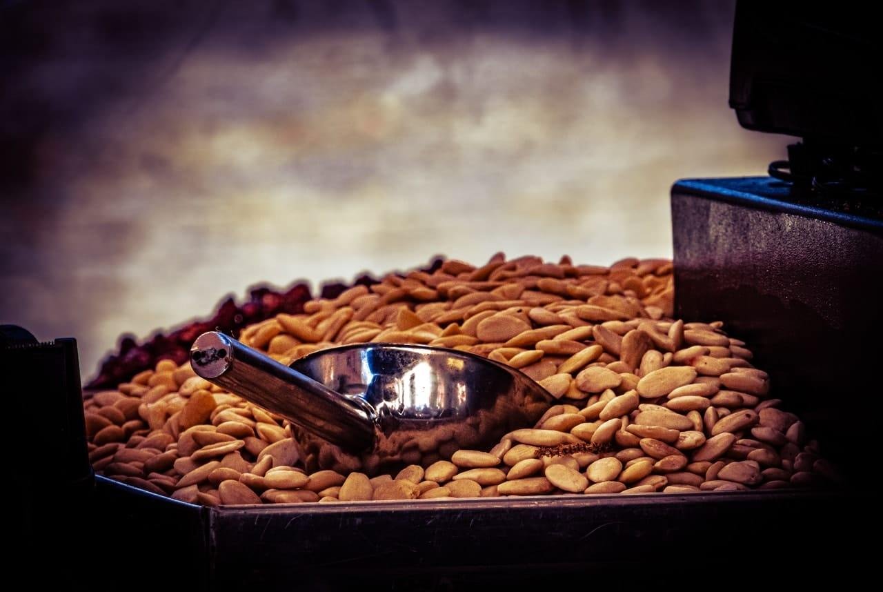 Metal scoop resting in a large bin filled with roasted nuts, with additional nuts piled behind and dark machine edges framing the left and right sides.