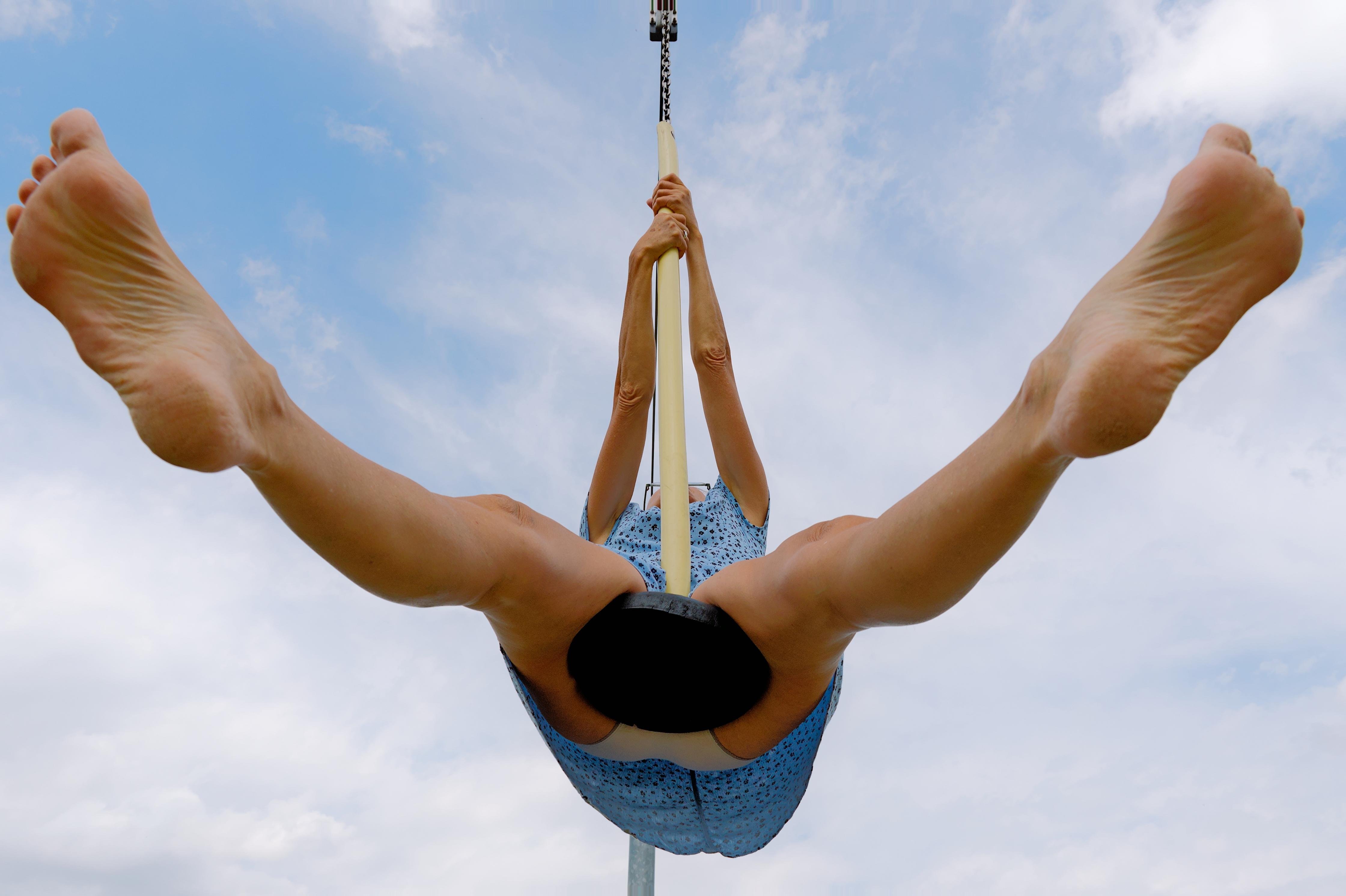 Person sitting on a swing, gripping the ropes above their head, viewed from below. Wearing a blue patterned garment, with legs extended outward against a backdrop of a partly cloudy sky. 
### 
Eine Person sitzt auf einer Schaukel und hält sich an den Seilen über ihrem Kopf fest (von unten betrachtet). Sie trägt ein blau gemustertes Kleidungsstück und hat die Beine nach außen ausgestreckt, vor dem Hintergrund eines teilweise bewölkten Himmels. 
