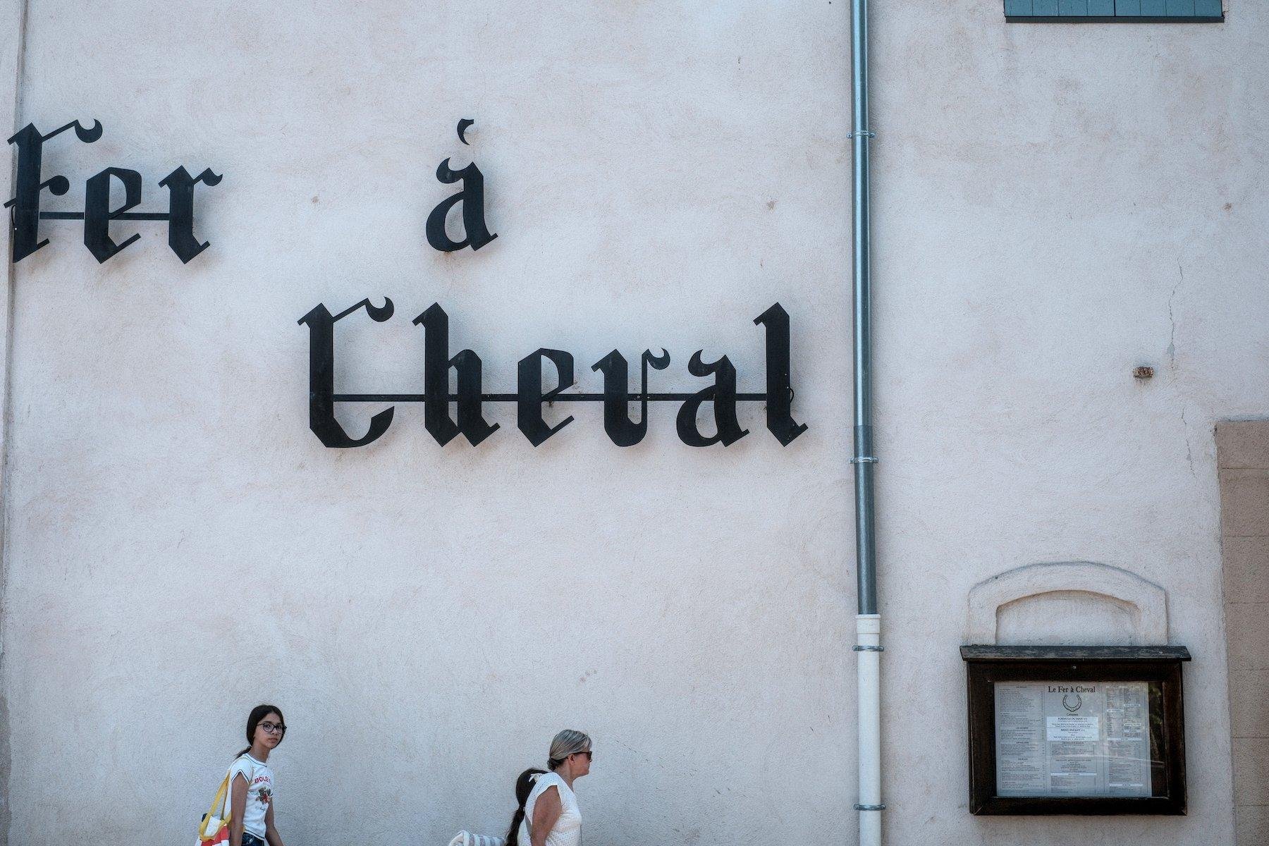 Photo horizontale. Un mur blanc sur lequel se trouve une évacuation de gouttière verticale, en fer sur la partie haute qui entre dans une partie blanche se chargeant de finir dans le sol. À gauche, une grande enseigne marquant le nom du restaurant, « Fer à Cheval » dans une typo ancienne. À droite du tuyau, le menu du restaurant. Des gens passent devant le mur, sous l’enseigne