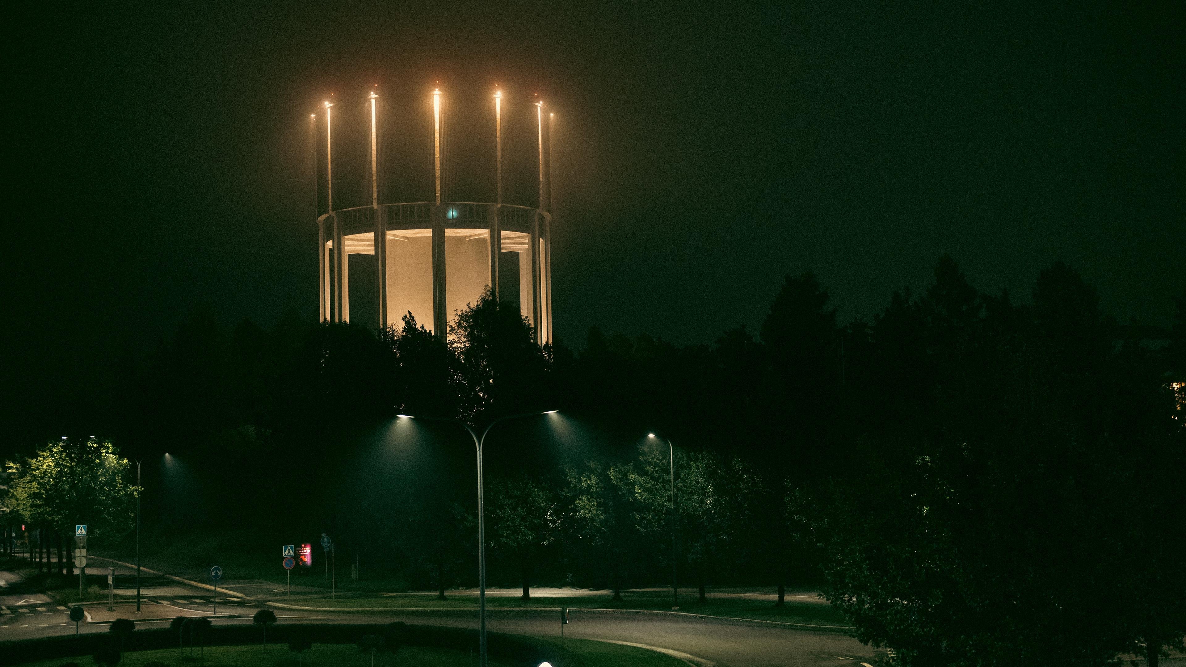 Circular water tower illuminated by vertical lights, surrounded by trees in a dark, misty night setting. Dim streetlights and a winding road visible in the foreground, creating a contrast between the well-lit structure and shadowy surroundings. Lappeenrannan vesitorni.