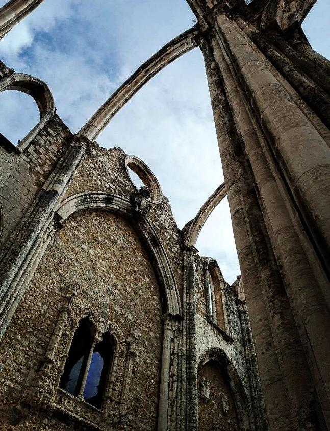This image shows the ruined arches and walls of a historical structure. The architecture appears gothic, with large stone columns and arched windows against a blue, cloudy sky. It might be the remains of an old church or monastery.