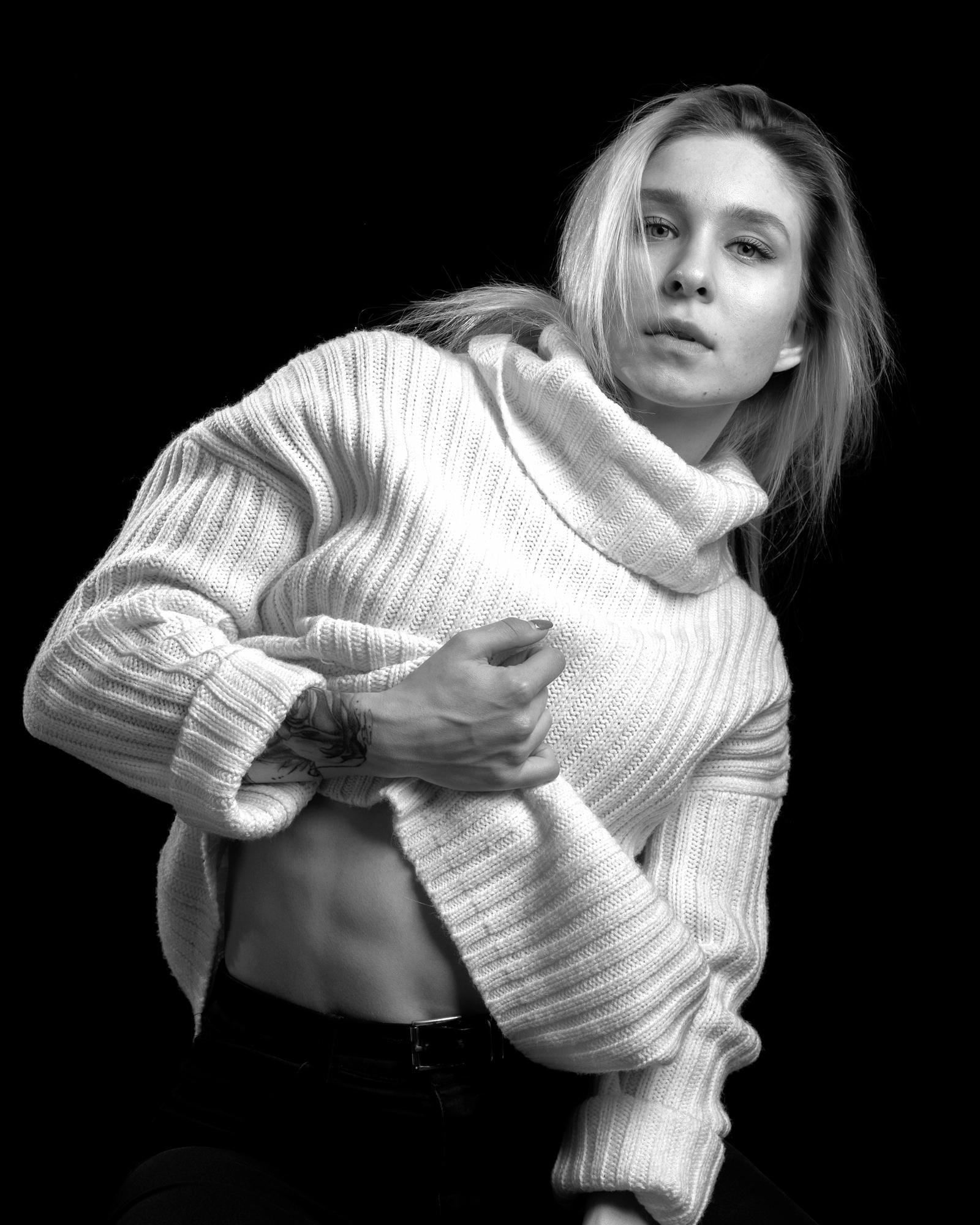 Young person with long hair wearing a white turtleneck sweater lifts it to reveal a toned midsection. The image is in black and white, highlighting the texture of the sweater and the person's tattoo on the left forearm. The background is solid black, emphasizing the subject.