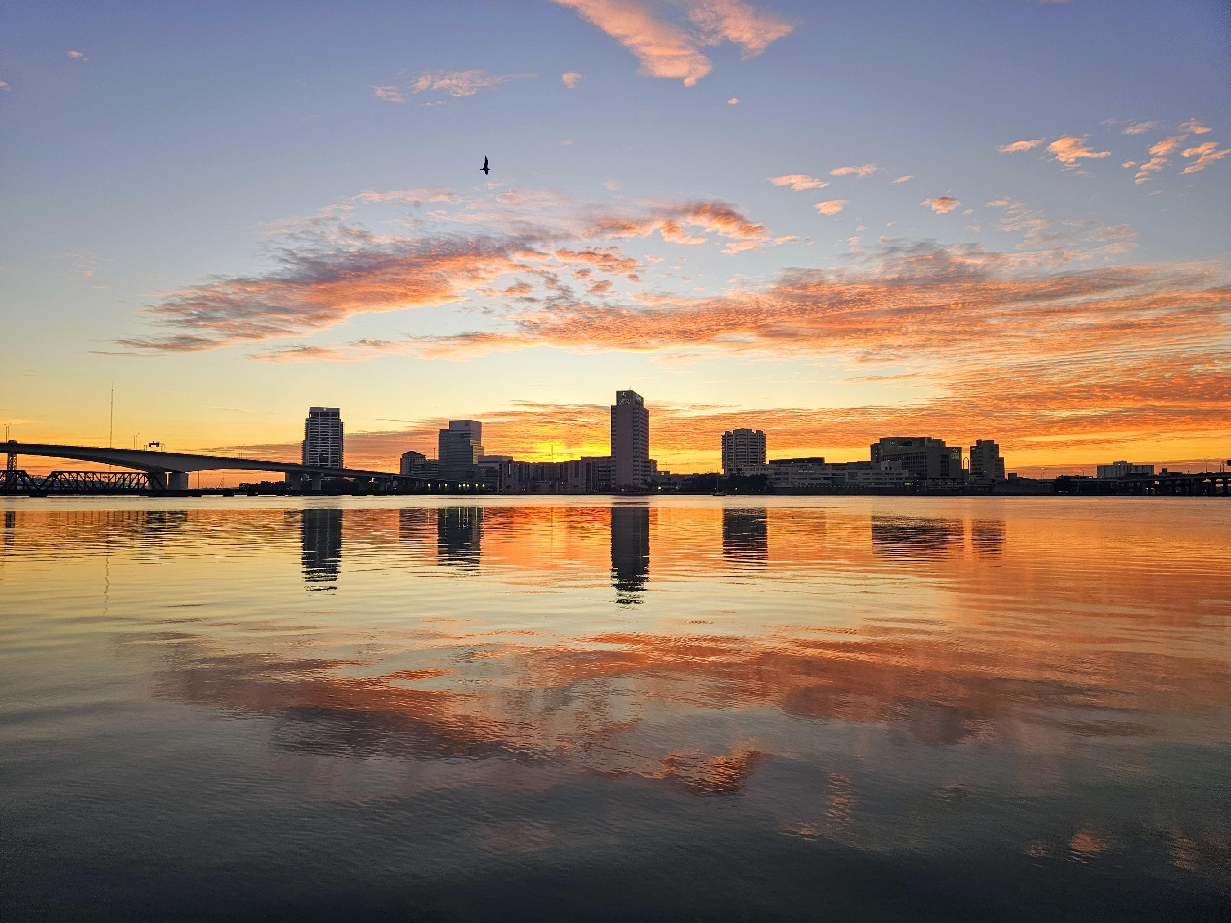 City skyline silhouetted against a vibrant orange and blue sunset, with clouds scattered across the sky. A bridge extends from the left, and a bird is flying near the top. Reflections of the skyline and sunset colors are mirrored on the calm water below.