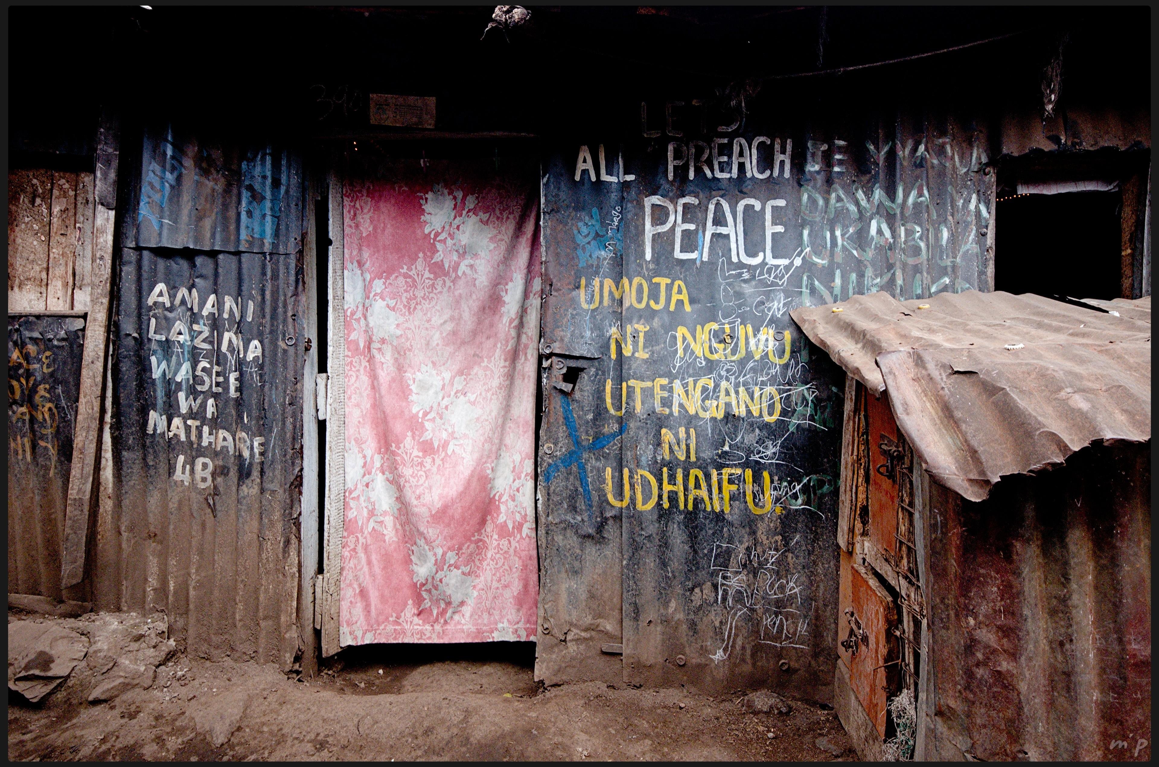 A horizontal colour photograph of the façade of a corrugated-sheet house in the heart of one of Nairobi’s slums. White and yellow inscriptions, in English and Kiswahili, cover the corrugated sheets, calling for peace. A very faded red fabric with white patterns serves as the front door. The uneven floor is made of compacted earth. To the right, a rabbit hutch, also made of corrugated metal sheets, can be seen.

///

Photo couleur horizontale d'une façade d'habitation en tôle au cœur d'un des bidonvilles de Nairobi. Des inscriptions blanches et jaunes, en anglais et kiswahili, couvrent les tôles ondulées, appelant à la paix. Un tissus rouge très délavé avec des motifs blanc sert de porte d'entrée. Le sol inégal est de terre battue. On distingue à droite un clapier, lui-même en tôles ondulées.