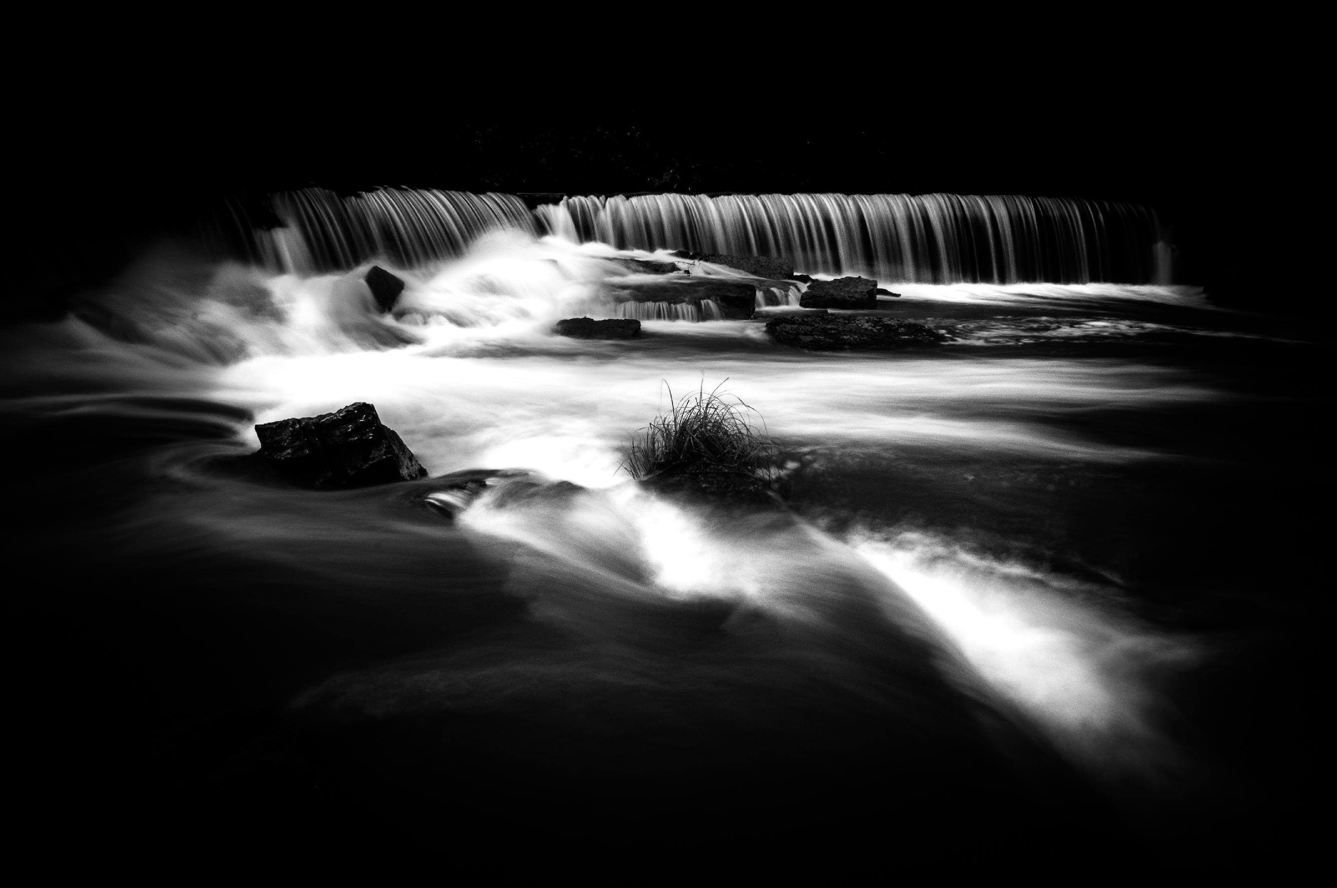 Black-and-white long-exposure photo of a low waterfall spanning the upper frame, with blurred water flowing over rocks into a river; a small clump of grass and several dark rocks sit in the foreground amid smooth, streaking currents.