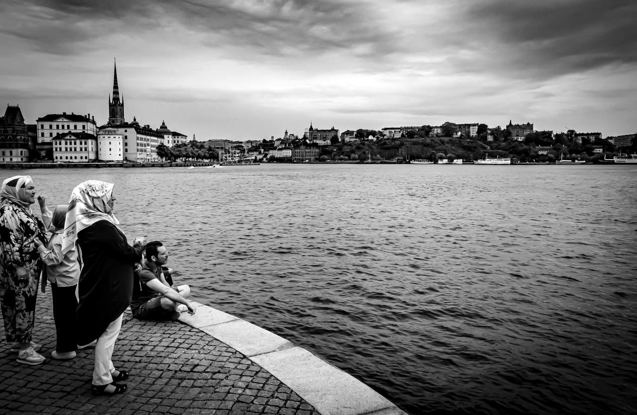 People stand and sit along a stone-paved waterfront, looking across a large body of water. The opposite shore features a town with historic buildings and a prominent church spire. The sky is overcast, creating a dramatic atmosphere. The image is in black and white, emphasizing contrasts and textures.