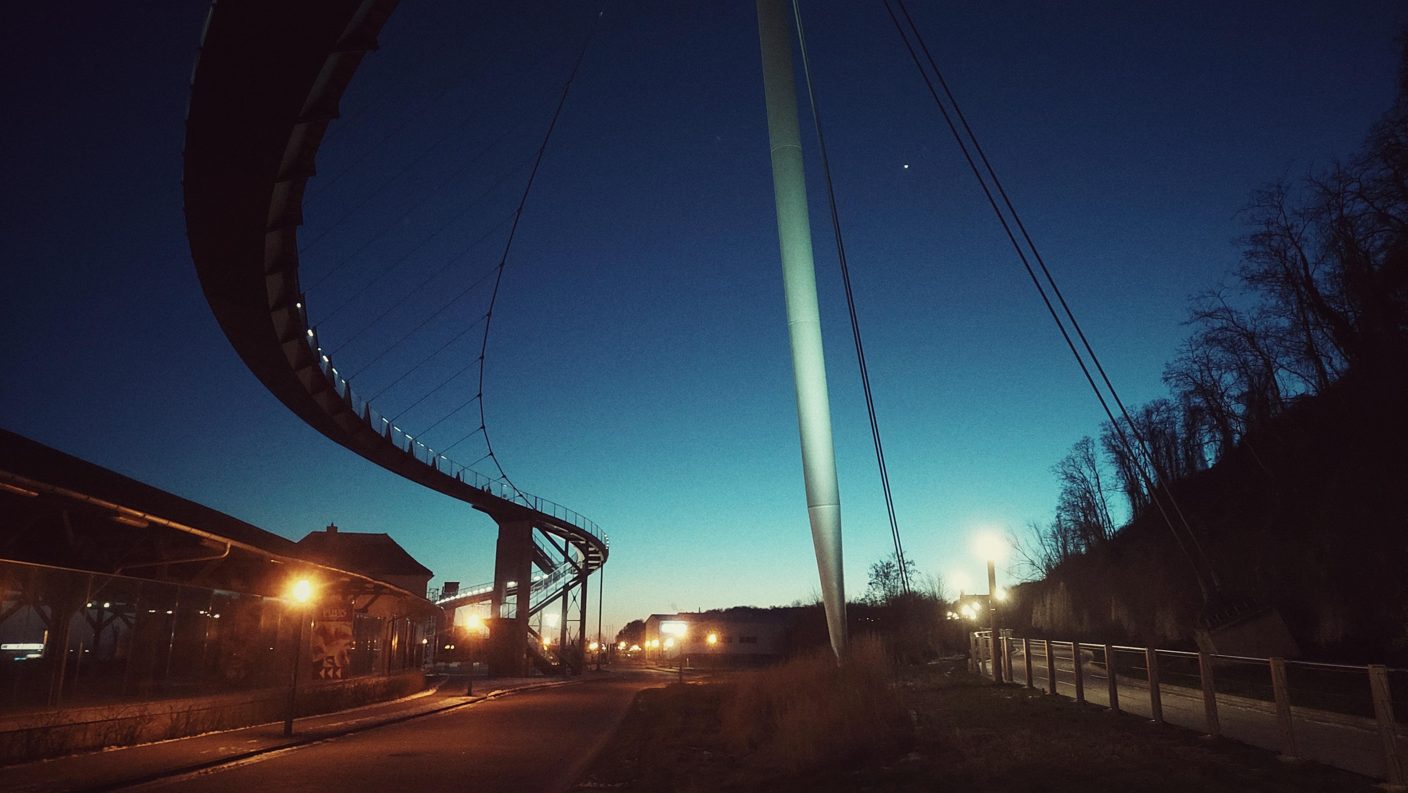 The image depicts a nighttime scene with a modern, curving bridge or elevated path extending across the scene. Streetlights illuminate the pathway below, casting warm glows. The sky is clear with a gradient from dark to light blue, indicating dawn or dusk. There are silhouettes of trees on the right and some buildings or structures on the left.
