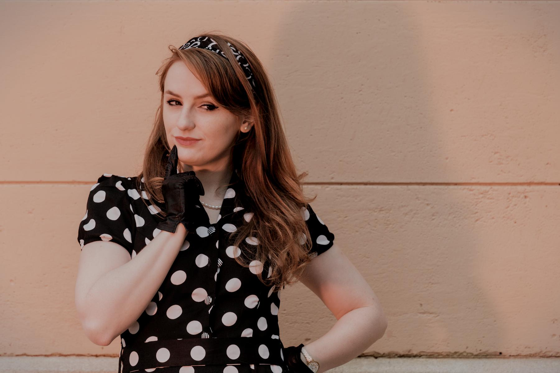 Photo horizontale. Plan taille d’une jeune femme rousse portant un bandeau dans les cheveux, une robe noire à pois blanc genre années 50 et des gants noirs en cuir. Elle porte son index droit à ses lèvres et le dos de sa main chauche est sur ses hanches. Elle a les yeux regardant en coin vers sa gauche avec un air mutin
