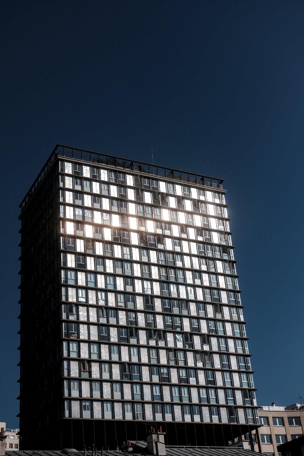 Photo verticale. Vue sur un immeuble à une distance respectable qui semble être posé sur plein de poteaux. Il est rectangulaire et ses multiples fenêtres réfléchissent le soleil, le faisant scintiller, sur fond de dégradé de ciel bleu