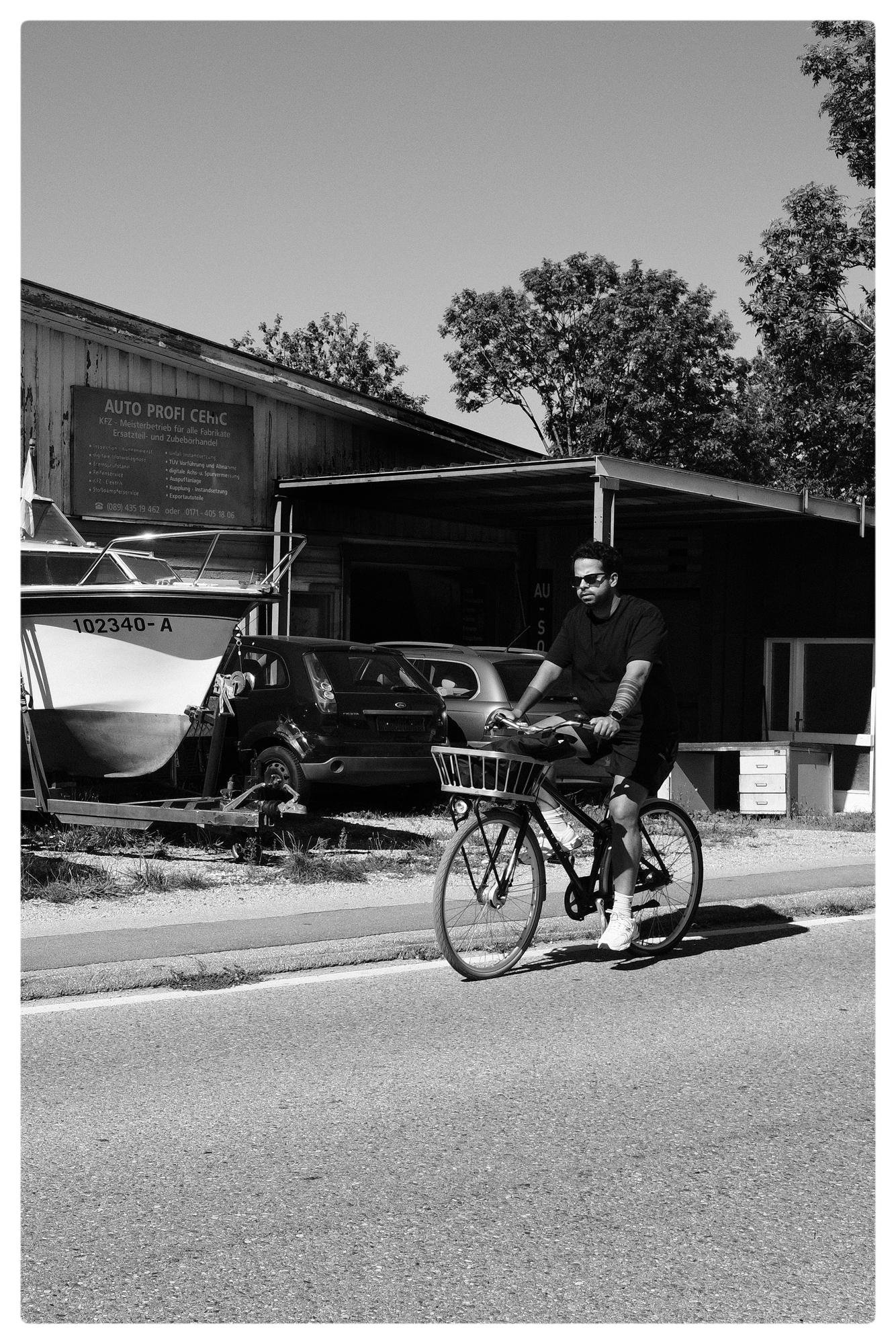 A person wearing sunglasses rides a bicycle with a front basket along a paved road. Parked cars and a boat on a trailer are visible in front of a wooden building with a sign labeled "Auto Profi CeHc". Trees in the background, clear sky overhead. The image is in black and white.