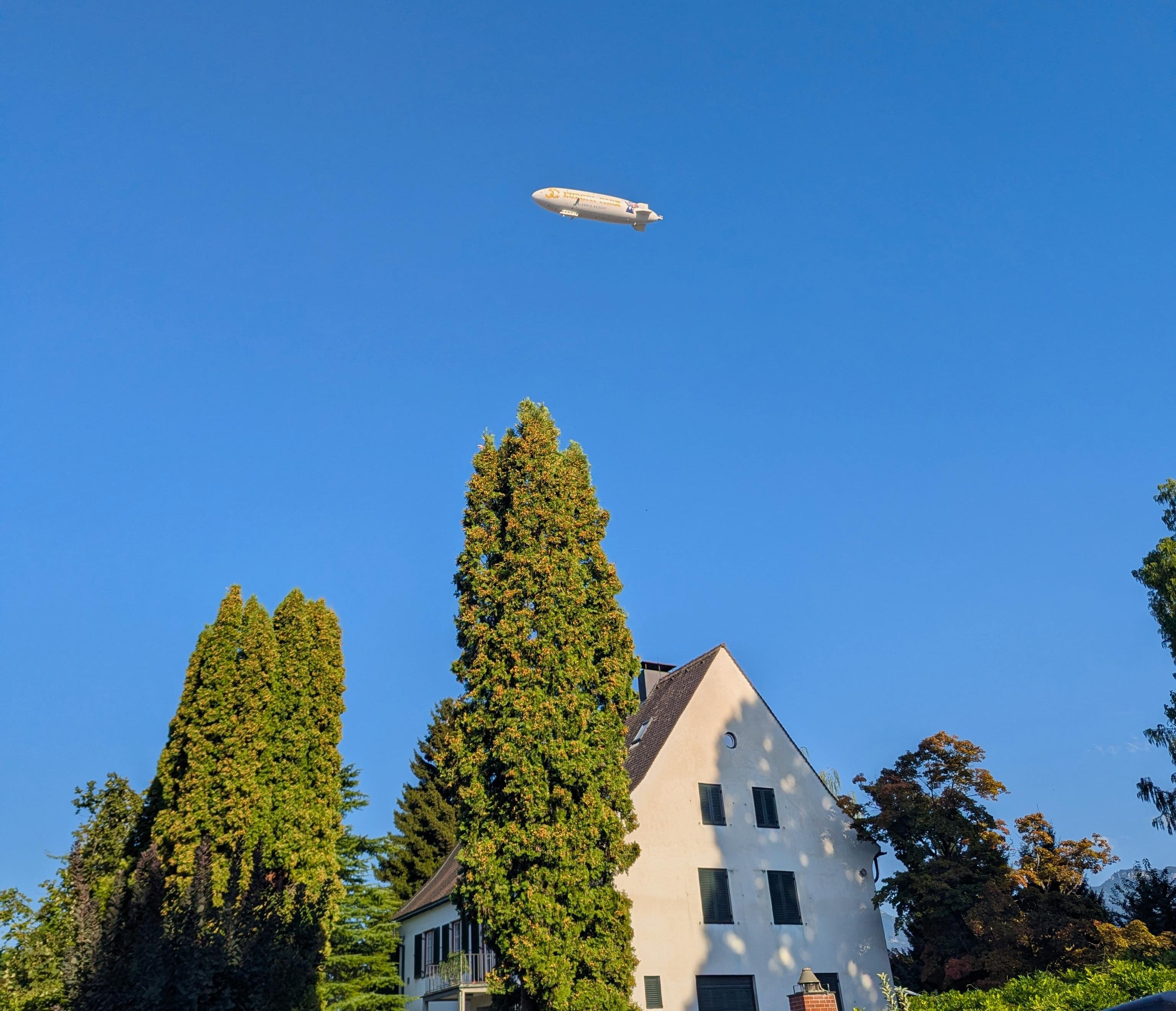 White blimp flying across a clear blue sky above tall evergreen trees and a white house with a steep roof.