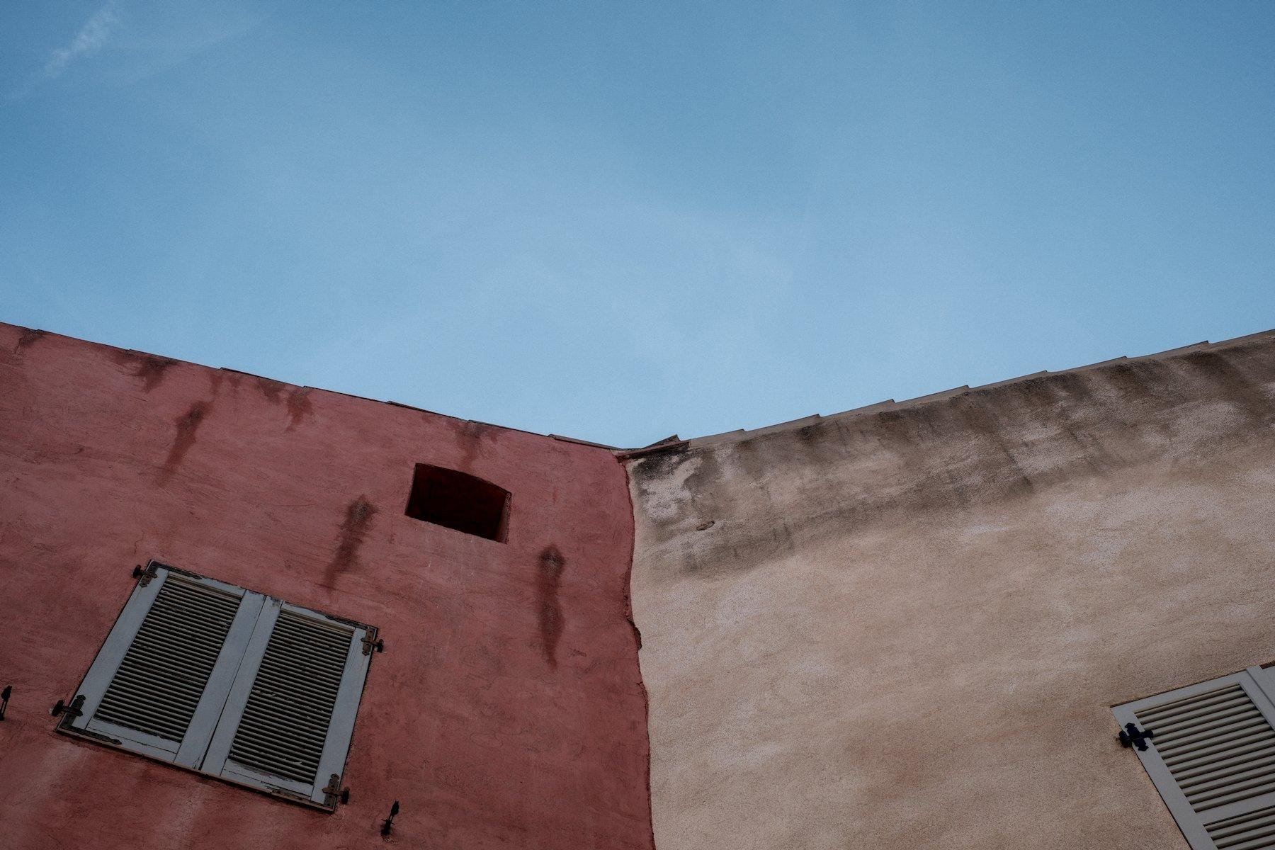 Photo horizontale. Contre plongée sur la façade de 2 bâtiments accolés mais de couleur différentes, ocre à gauche et beige à droite, avec le ciel bleu légèrement voilé de nuage en haut, divisant l’image en trois