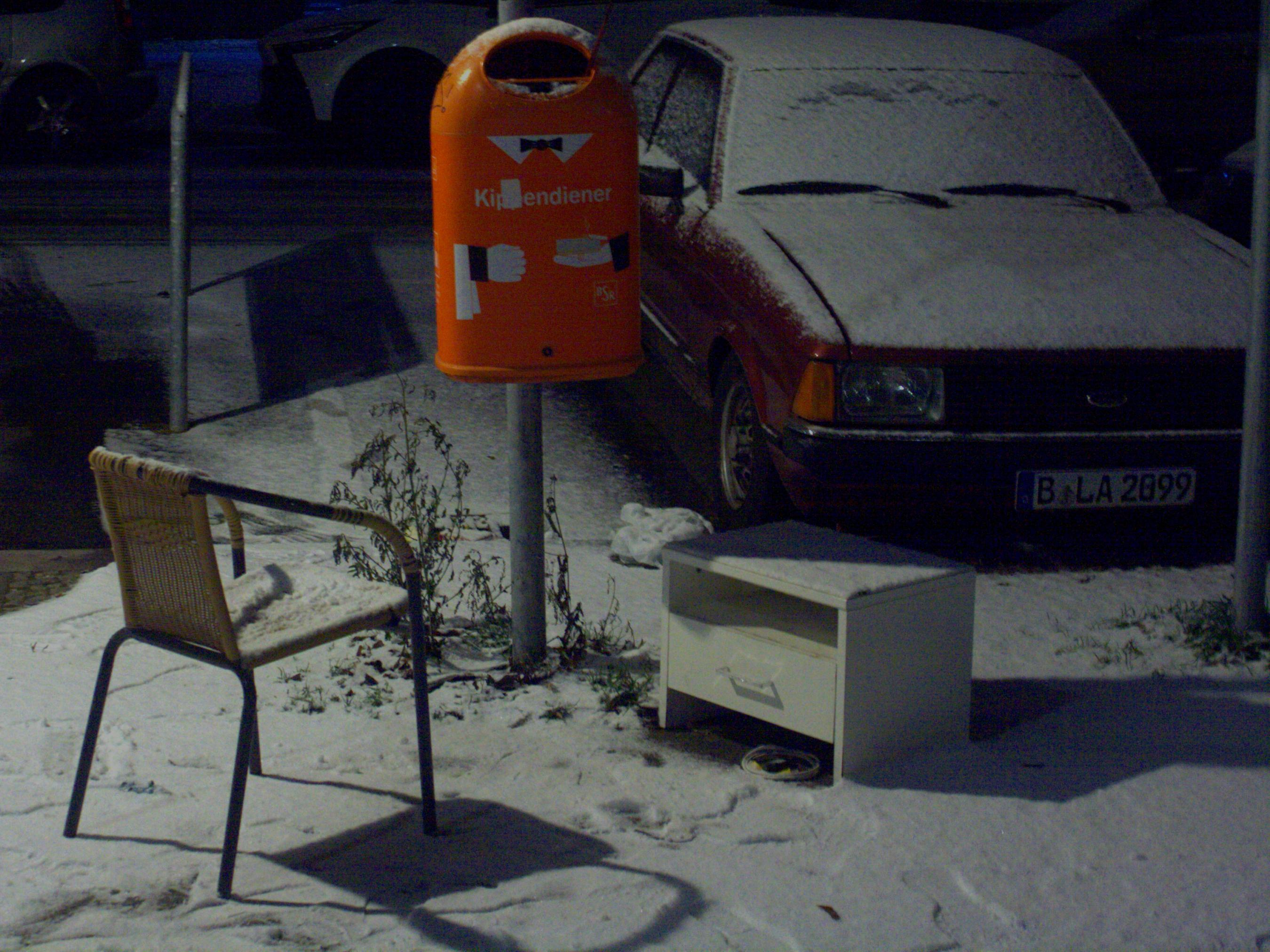 a photograph of a orange rubbish bin with a chair and night desk around it, everything is covered in snow