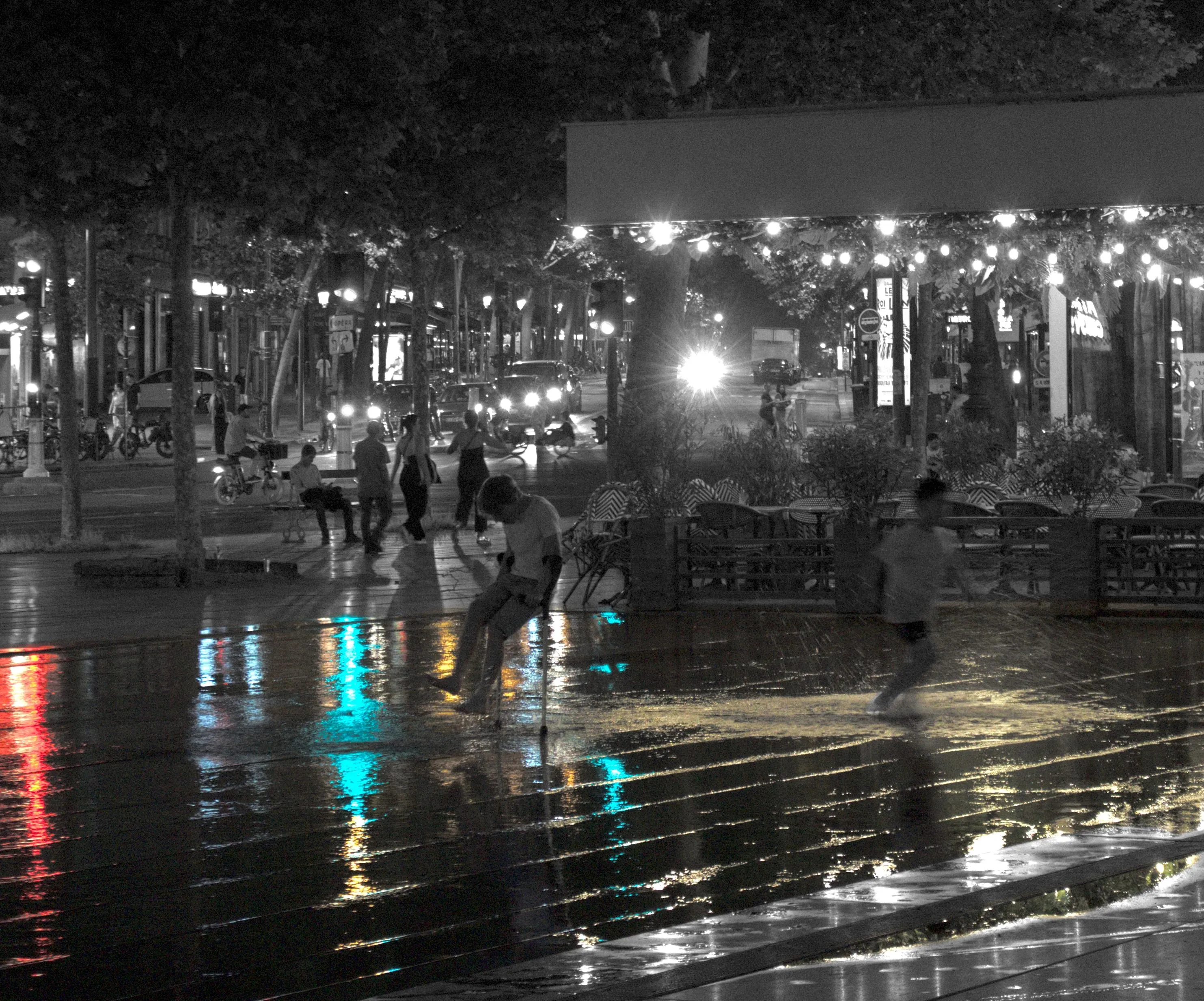 Photograph of children playing around on a public square. Most of the surface of the square visible in the image is covered in water. City lights are reflected on the water surface. One of the kids is using crutches and made a water splash. The other kid seems to be running away from the first. The surface of the water has a green coloration, reflecting the trees surrounding the square. The reflected city lights form bright red, yellow and blue columns on the water surface. The rest of the photograph is in monochrome, including the children that are detoured.