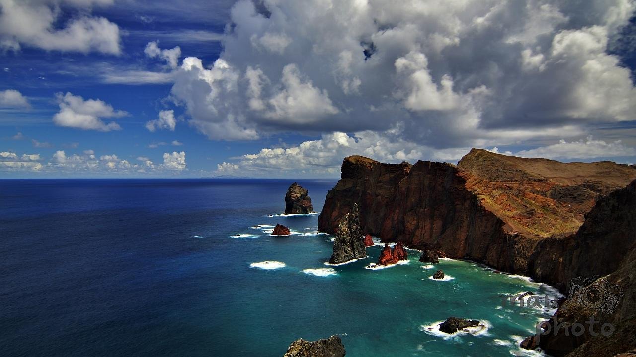 Rocky coastal cliffs with rugged formations extend along a deep blue ocean. Towering clouds dot the bright sky above, casting shadows on the sea. Bright sunlight illuminates the reddish-brown hues of the eroded rock face. Waves crash against the base of the cliffs, creating white frothy patterns on the water's surface.