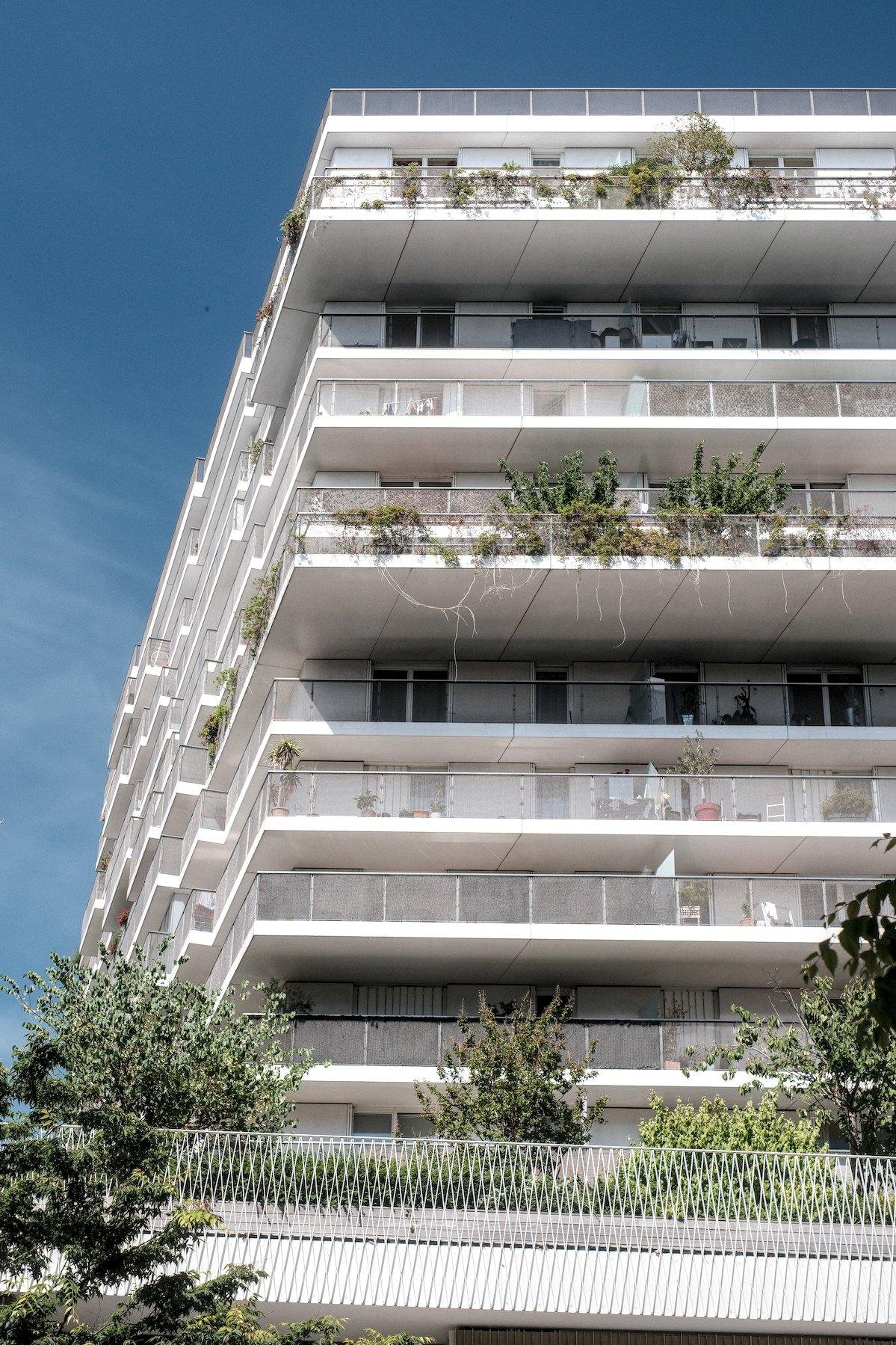 Photo verticale. Un immeuble blanc fortement éclairé par le soleil, dont on voit une face, et celle de gauche en perspective. Perspective qui donne un effet d’escalier aux balcons