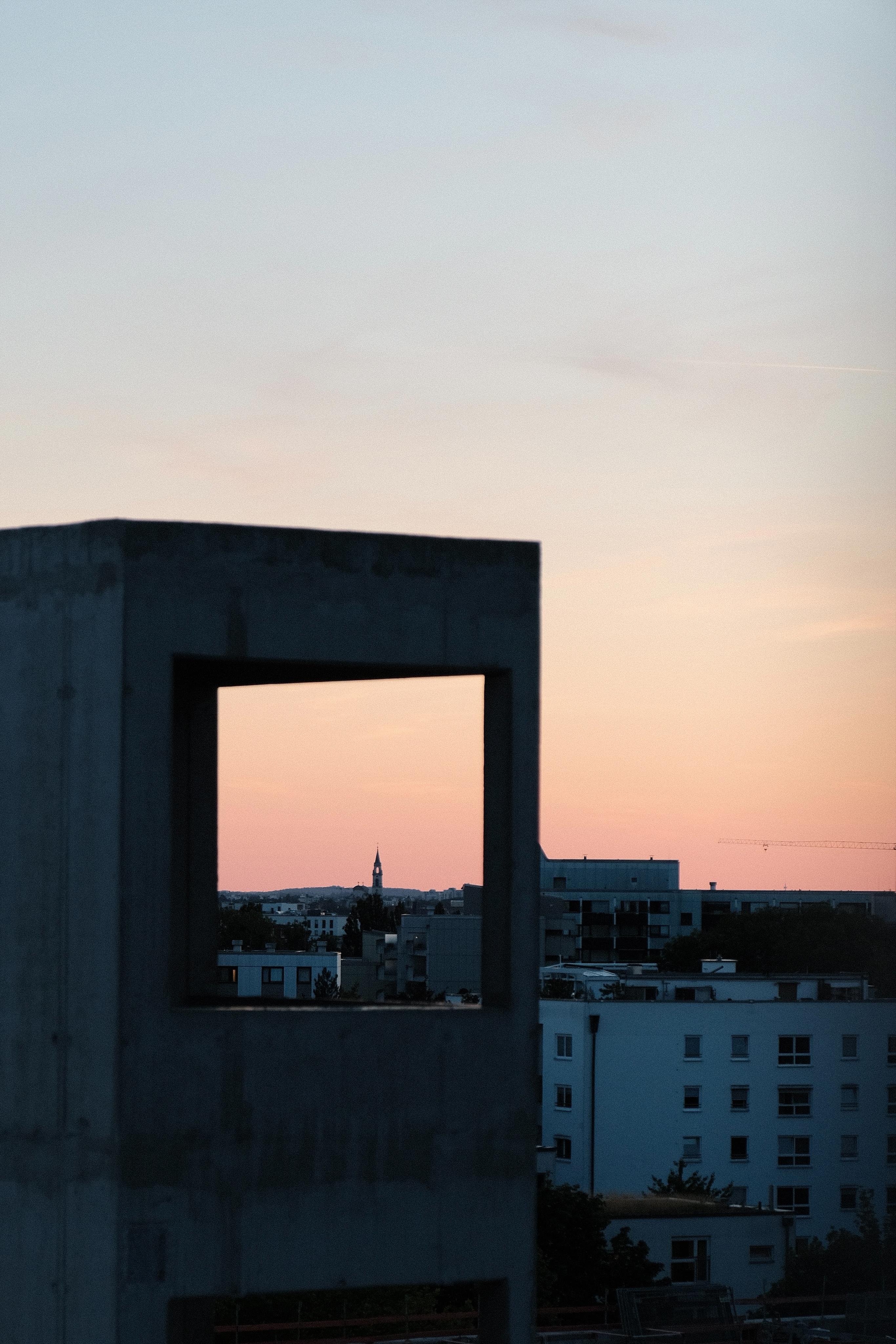 View from a rooftop terrace over a flat city skyline. In the foreground, an unfinished concrete building with a window frame frames a small church tower in the skyline. All is set in a red-yellow gradient sky shortly after sunset.